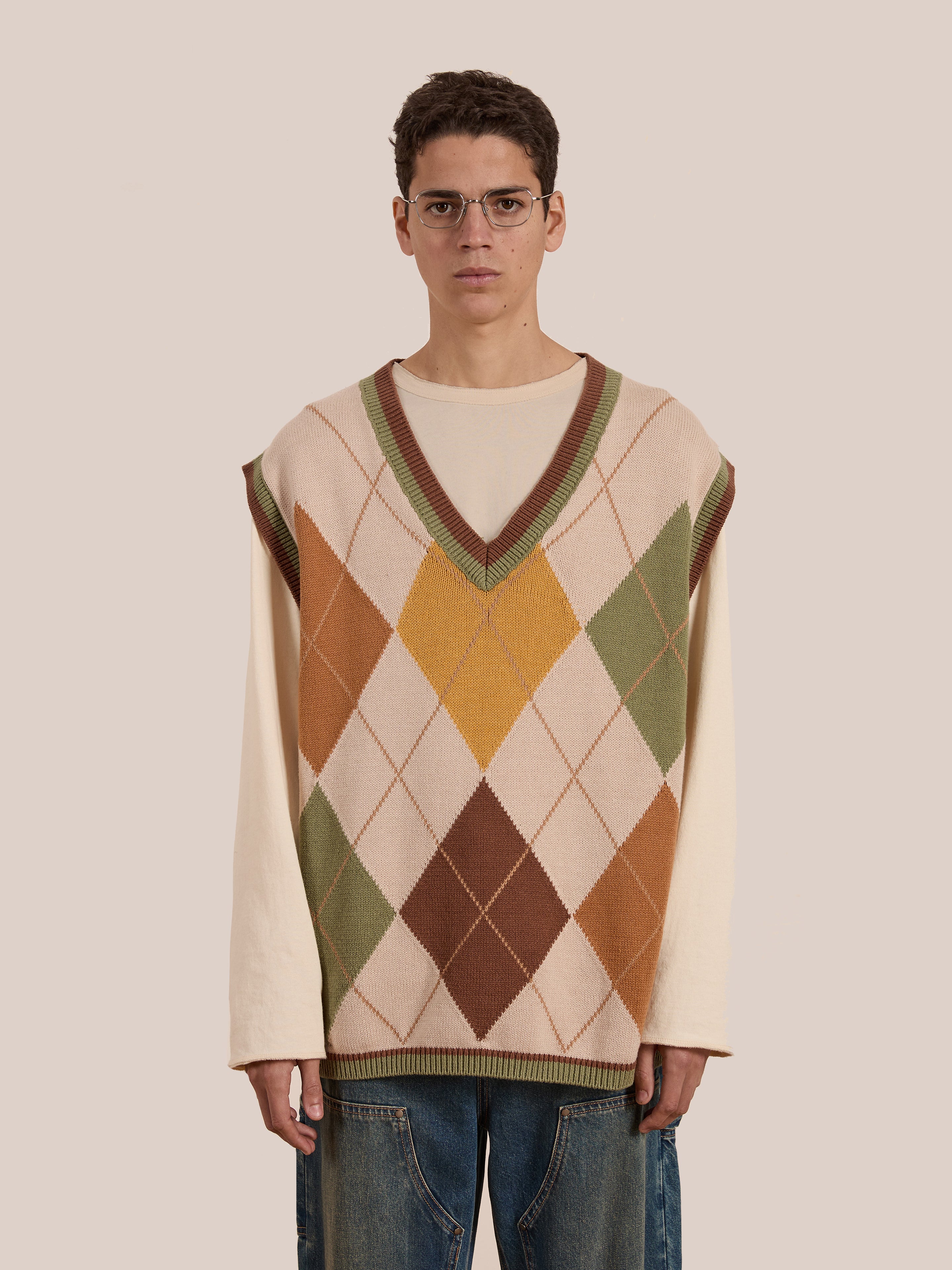 A person wearing glasses, a cream long-sleeve shirt, and an oversized, vintage-inspired Argyle Knit Vest by FOUND in various colors stands against a plain background.