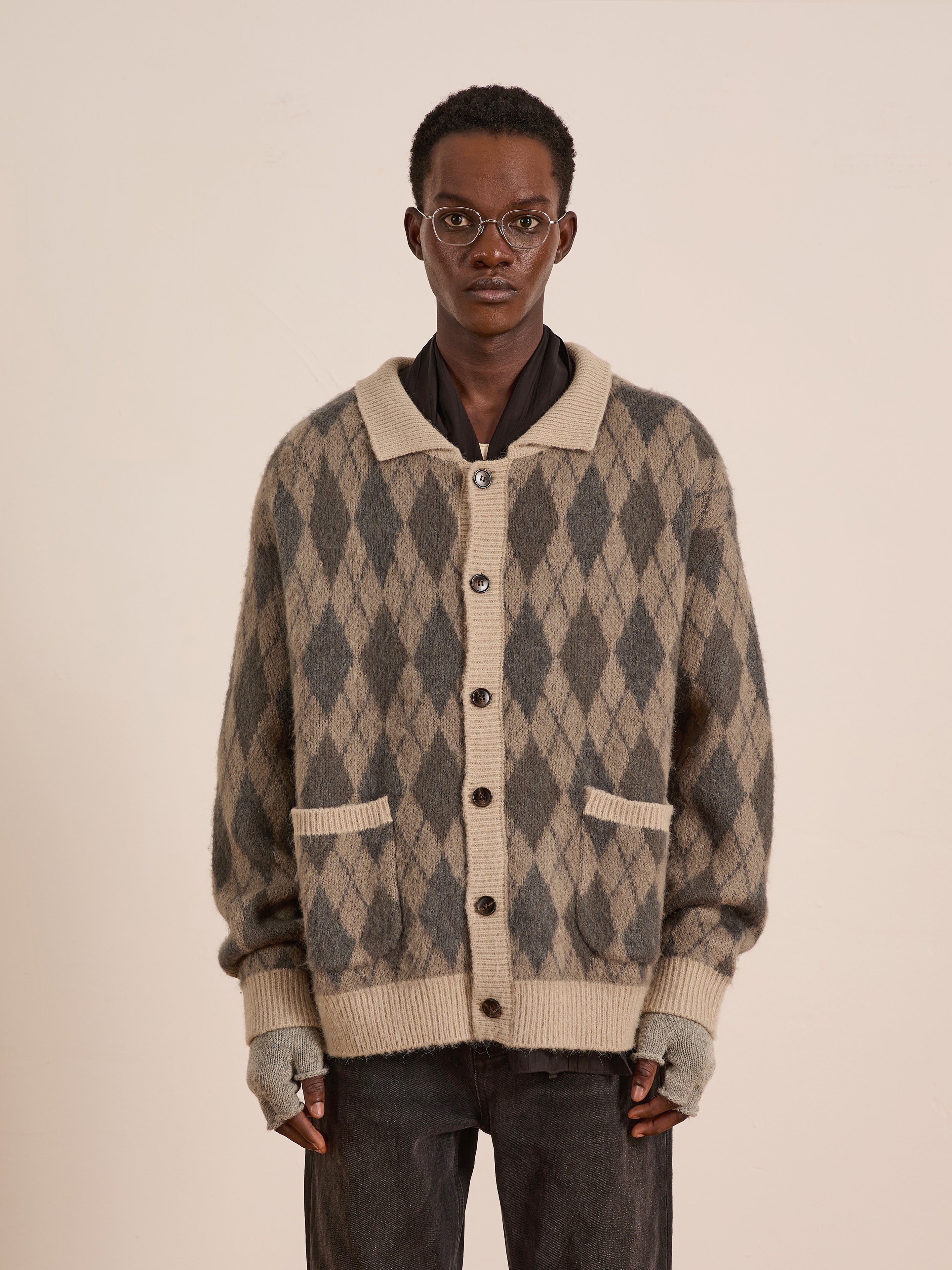 A person in glasses wears the FOUND Argyle Cardigan, a beige and grey wool blend with large buttons, relaxed-fit sleeves, and front pockets, standing against a plain light background.