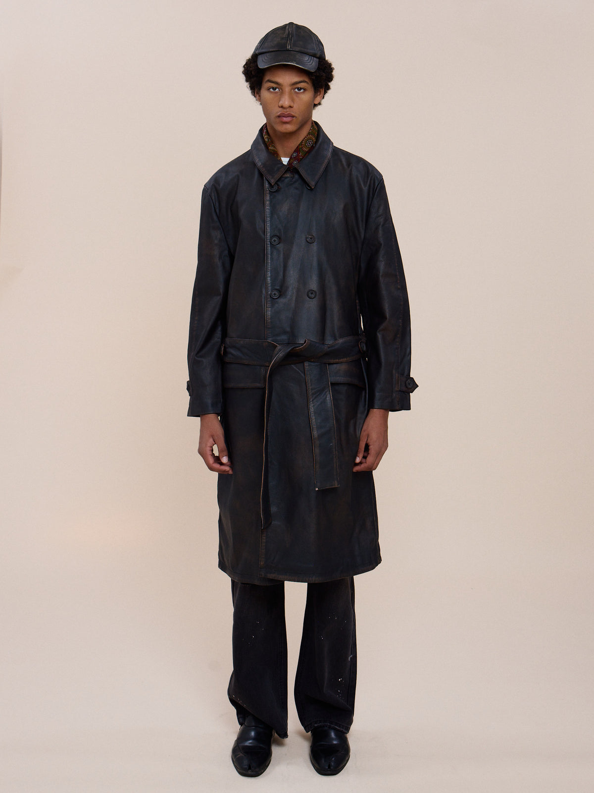 Person stands against a plain background in the FOUND Belted Leather Trench Coat, black pants, shoes, and matching cap, facing the camera with a neutral expression.