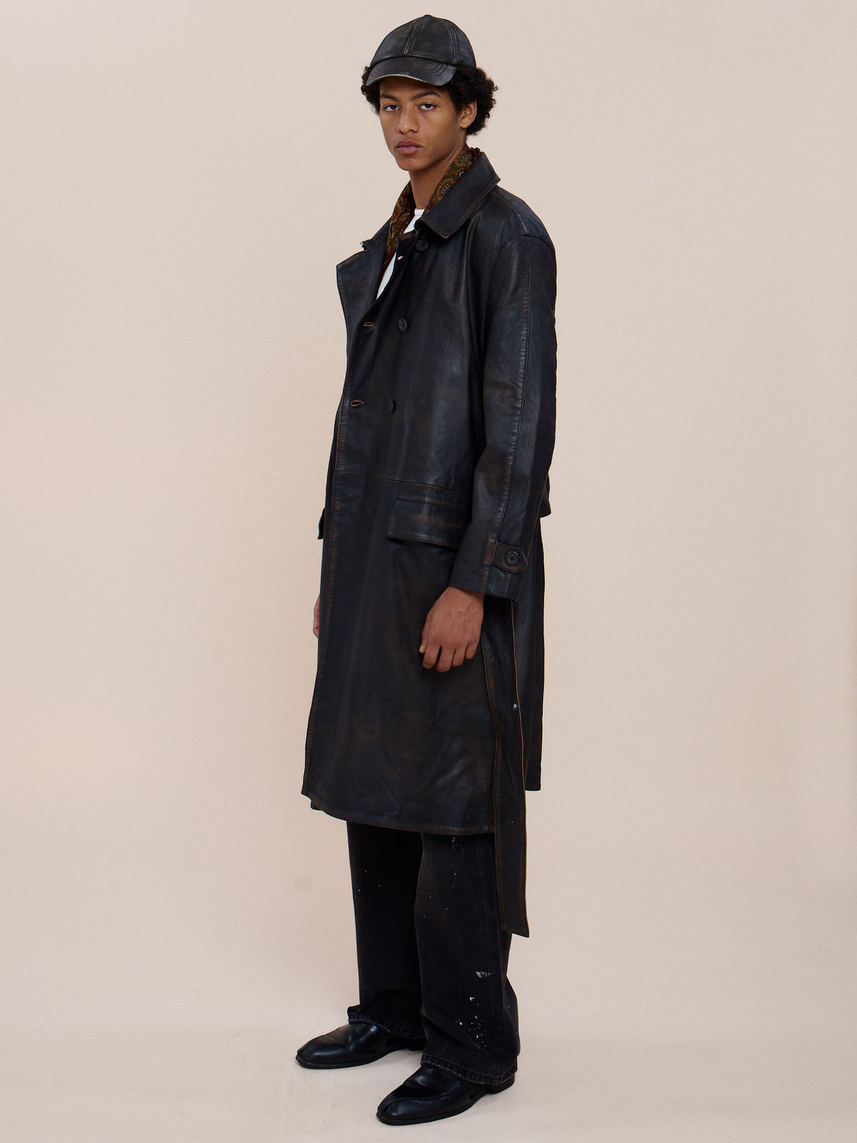 Person wearing the FOUND Belted Leather Trench Coat with black pants, black shoes, and a dark cap, standing against a plain beige background.