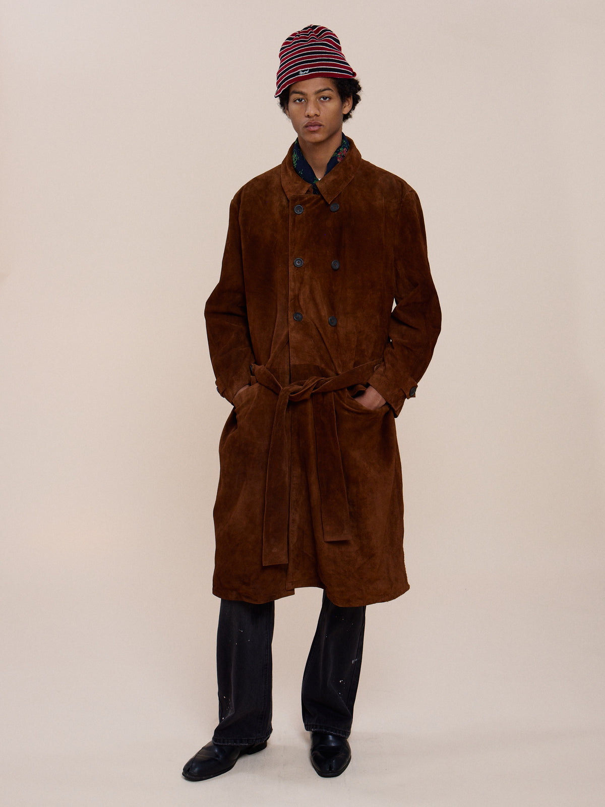 A person faces forward in the FOUND Belted Suede Trench Coat, paired with dark pants, black shoes, and a red-and-black striped hat against a plain light background.
