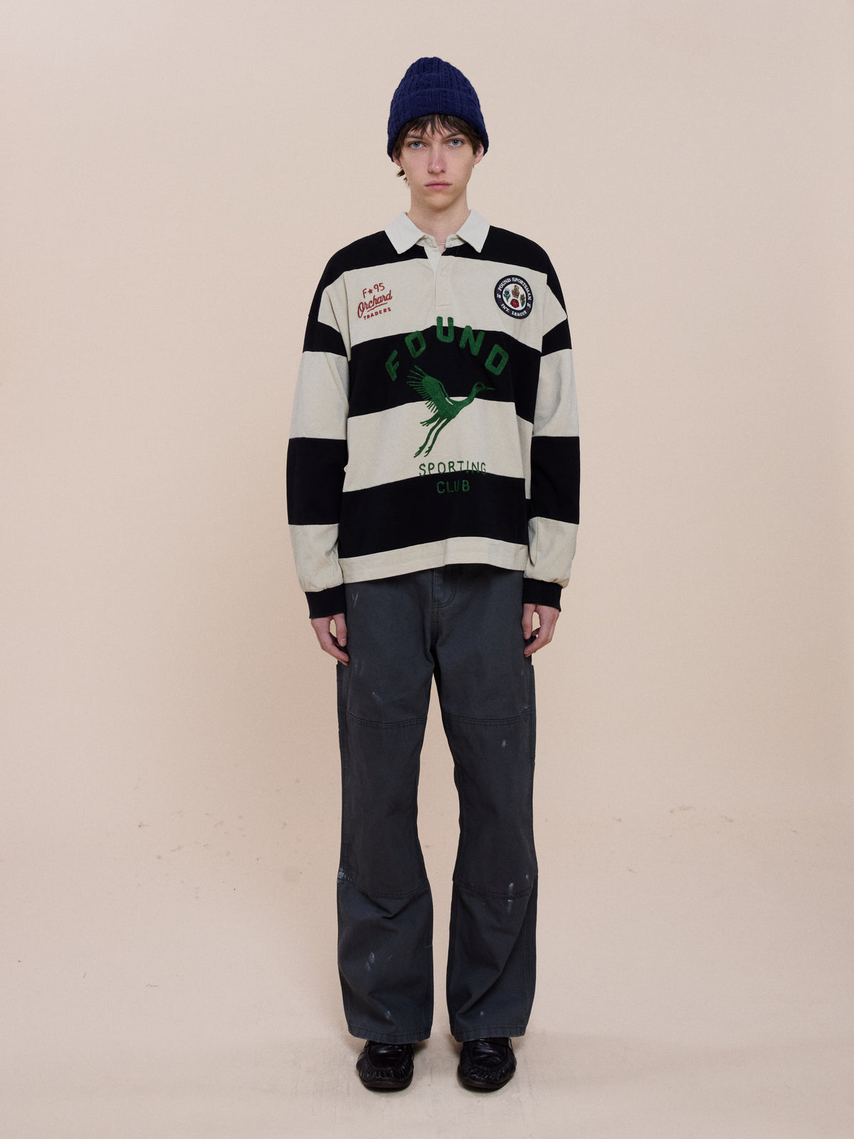 Person stands against a light background wearing the FOUND Bird Logo Rugby, loose dark pants, blue knit hat, and black shoes.