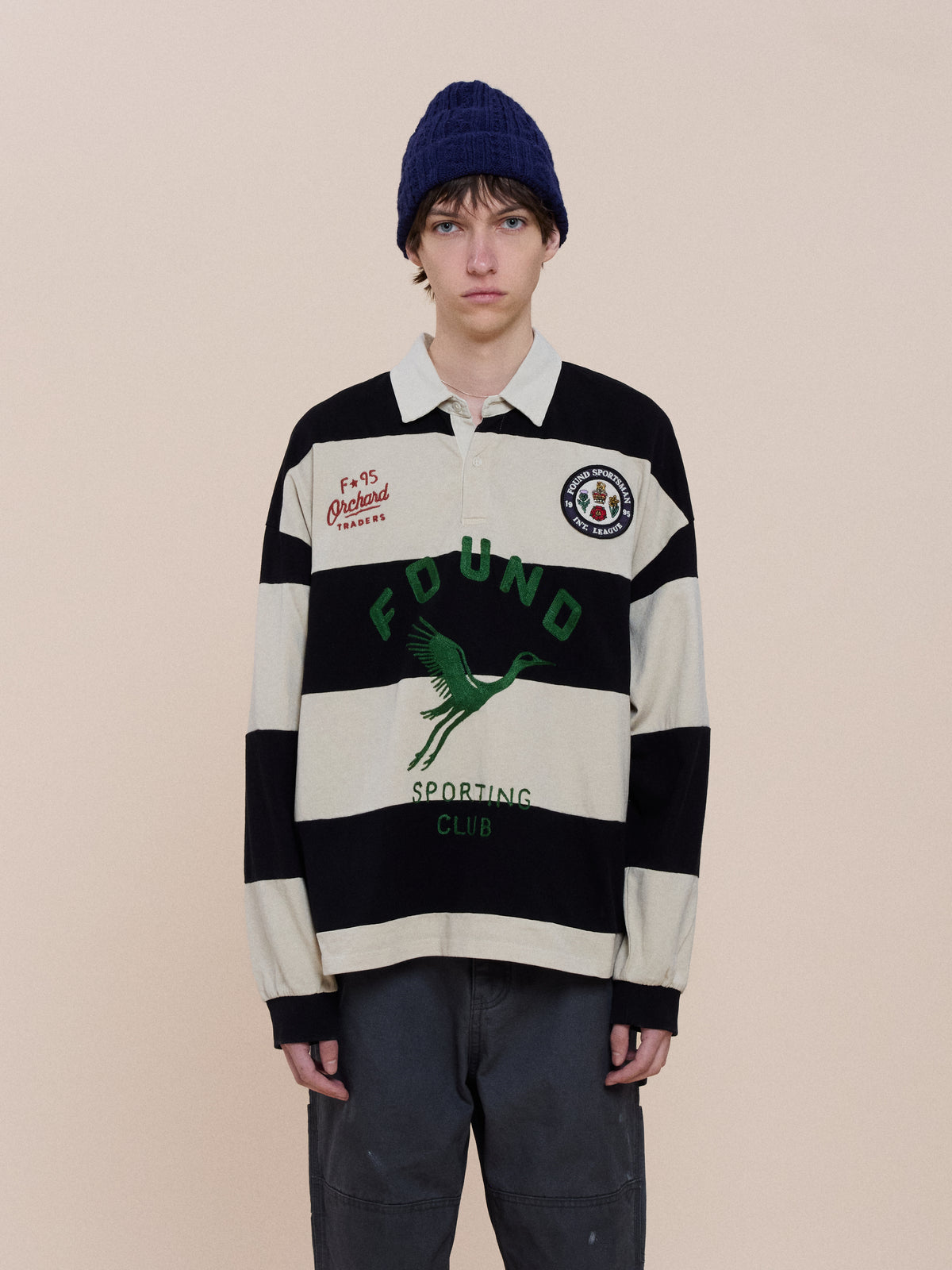 A person wears a navy knit beanie, FOUND Bird Logo Rugby, and dark pants, standing against a plain beige background.