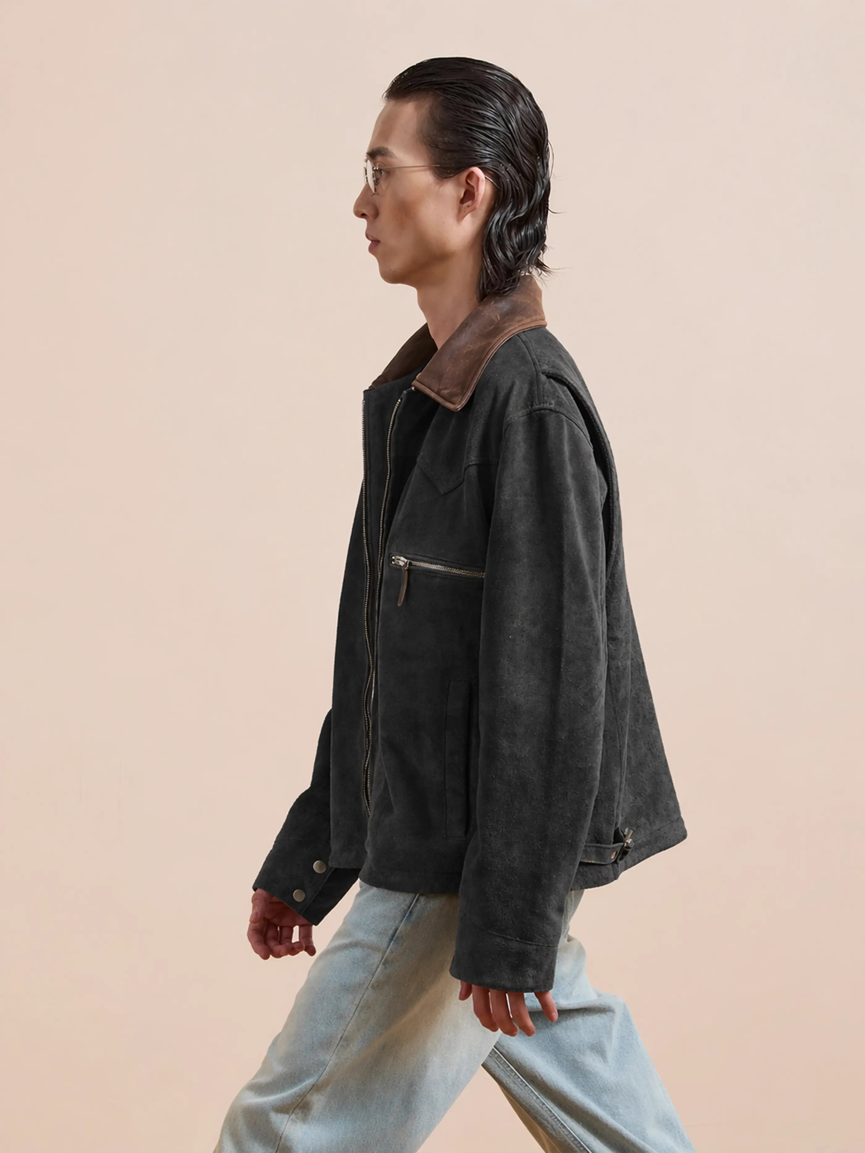 A person with slicked-back hair and glasses wears the FOUND Suede Western Work Jacket—a vintage black suede design with a brown collar—paired with light jeans, walking against a plain beige background.