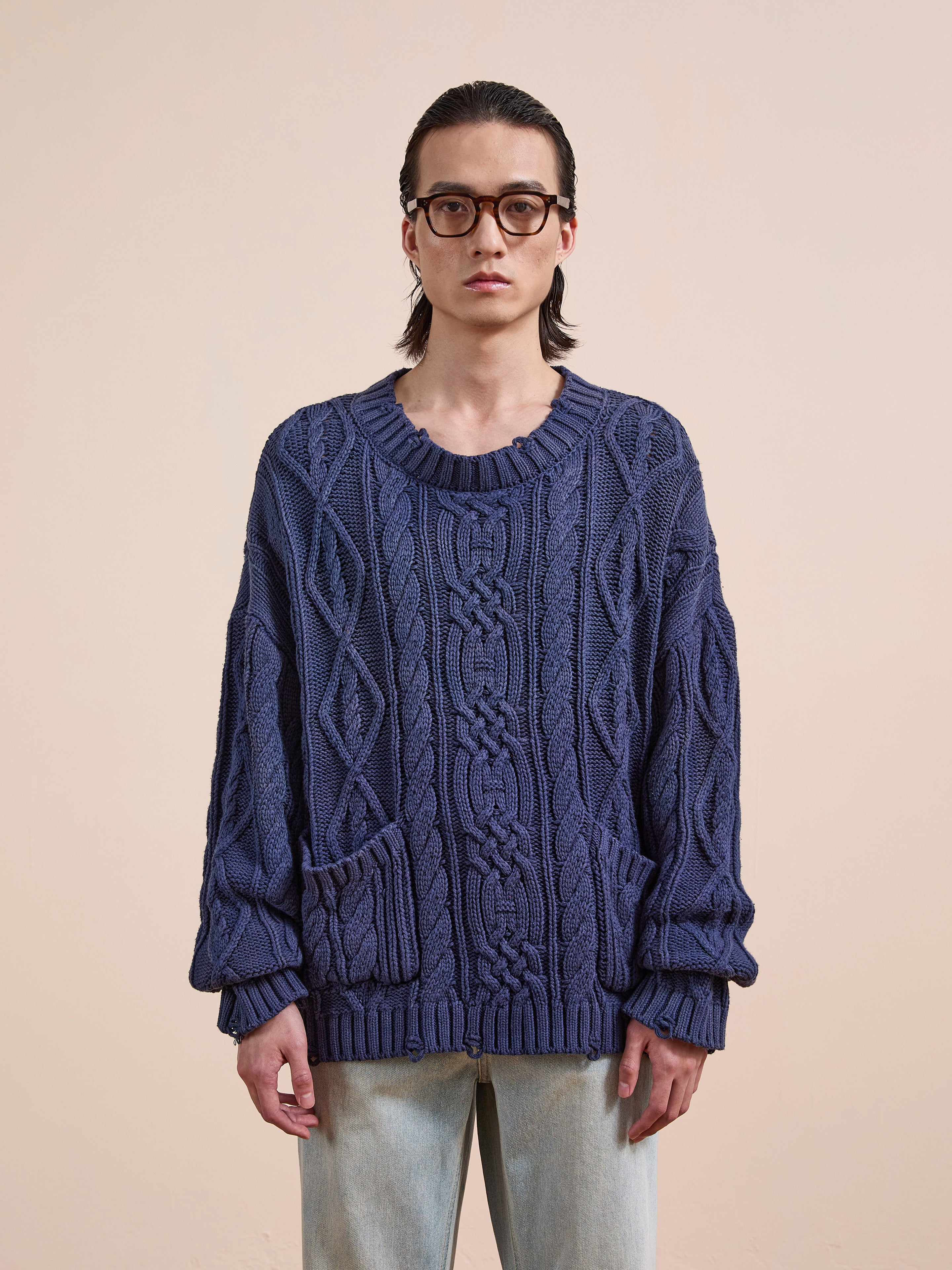 A person with long dark hair and glasses wears the Found Cable Knit Pullover Sweater in navy blue with pockets and light jeans, standing against a plain beige background.