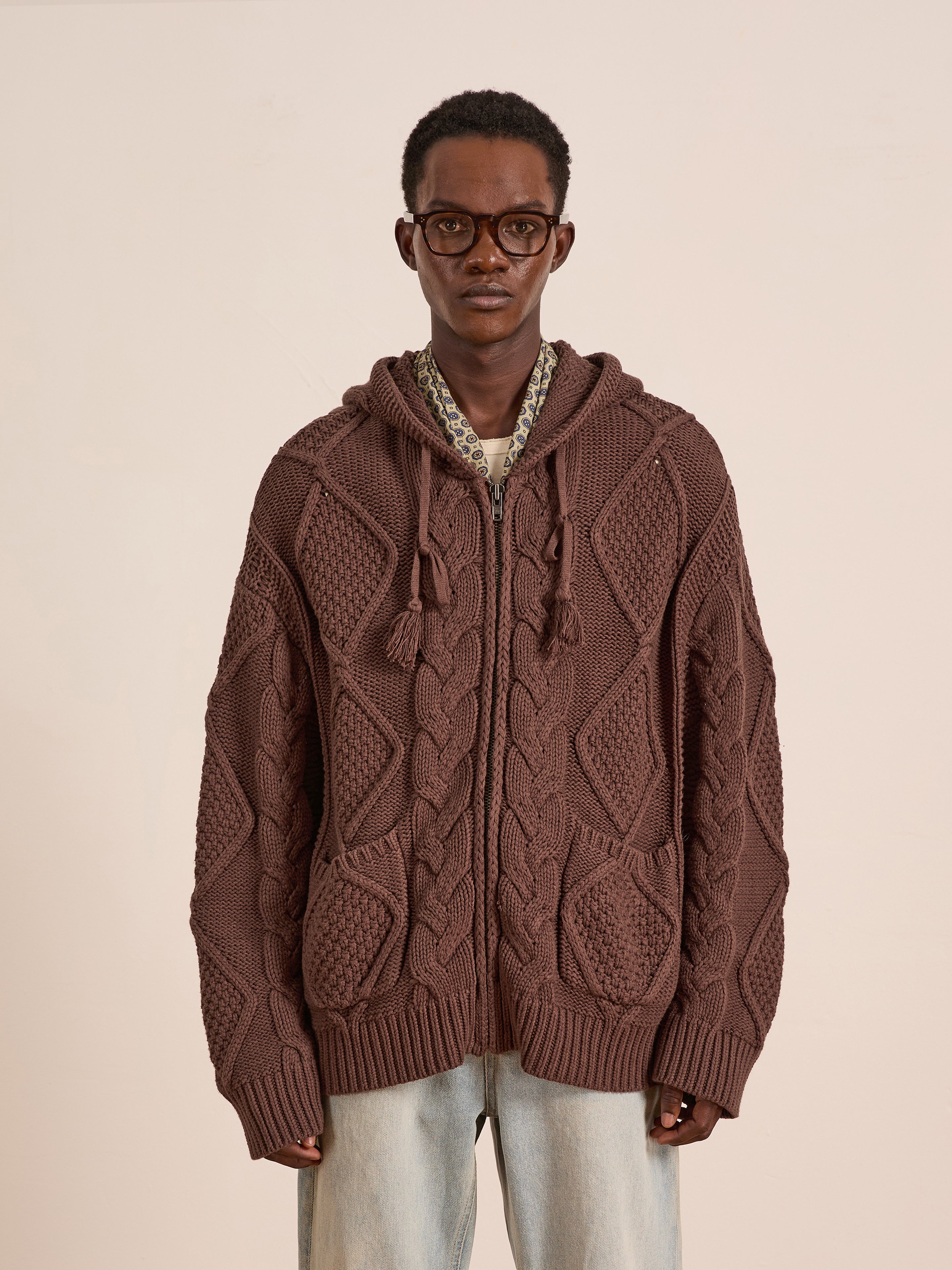 A person wearing glasses, light-wash jeans, and the FOUND Zip Up Cable Knit Hoodie stands facing the camera against a plain background.