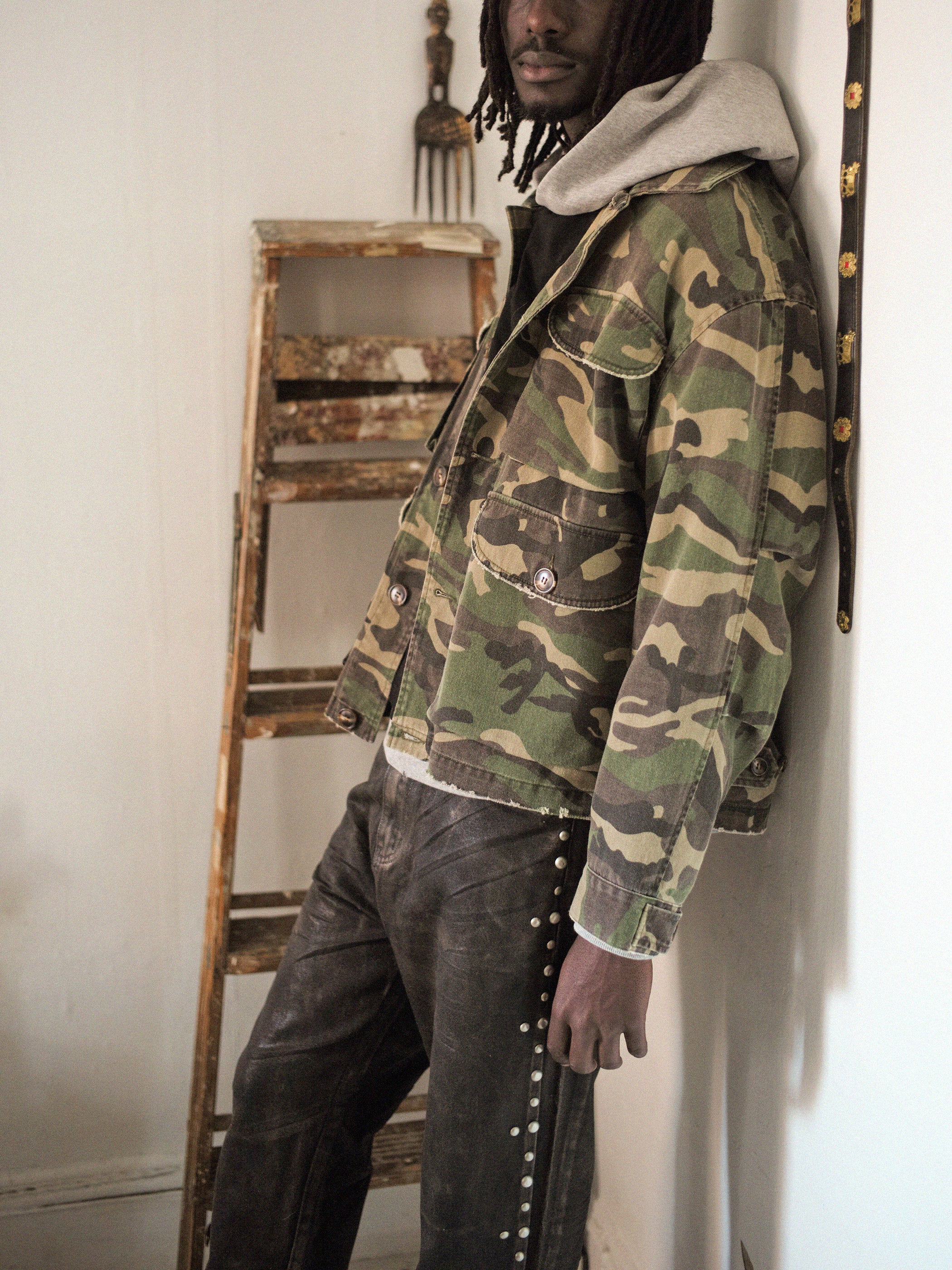 A person leans against a wall wearing the FOUND Camouflage Twill Utility Cargo Jacket and dark studded pants, with a wooden ladder nearby.