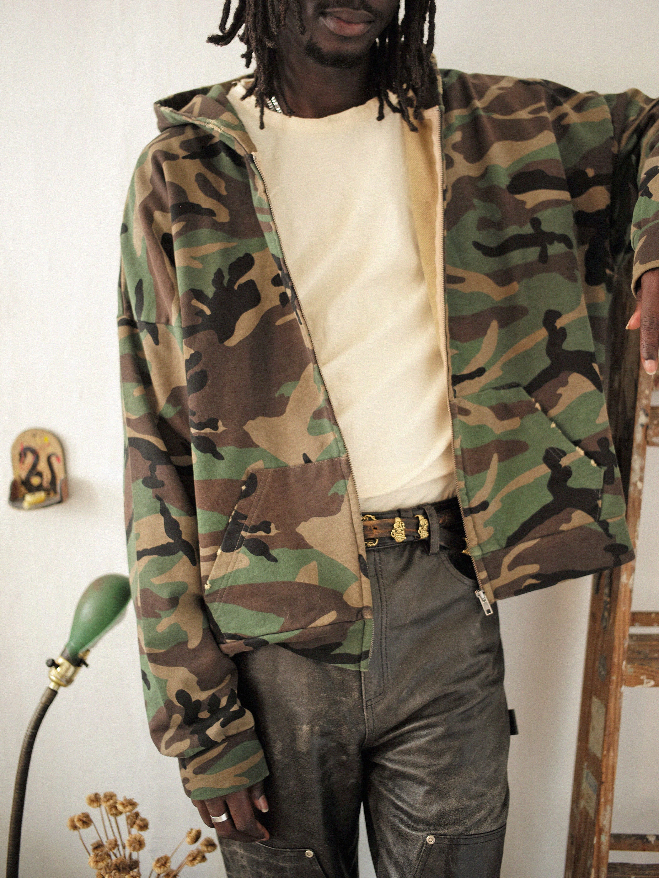 A person in a FOUND Washed Camouflage Zip Hoodie, white shirt, and black pants stands beside a ladder and a green lamp, with the camo print adding a vintage touch to their relaxed style.