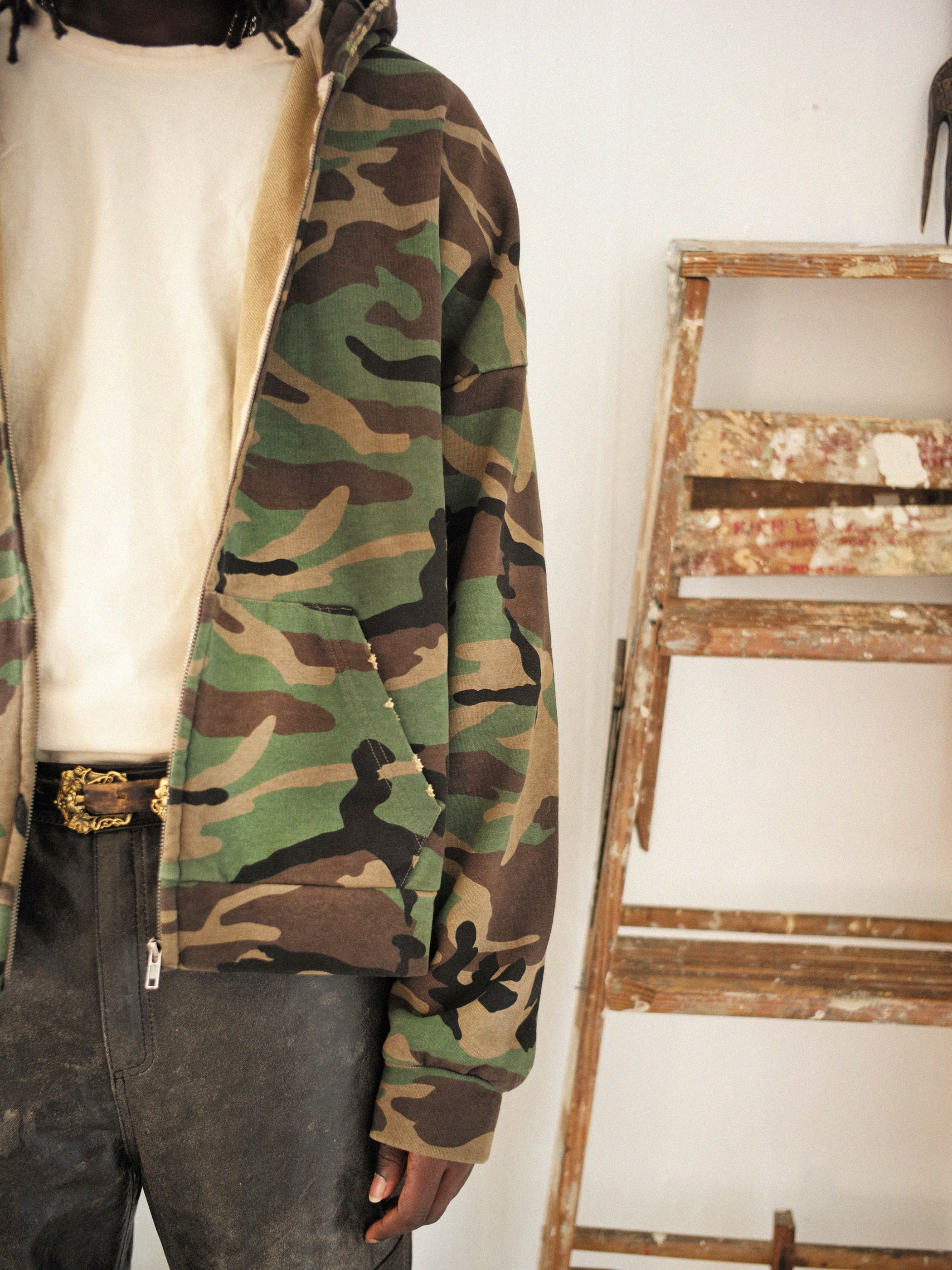 A person wearing the FOUND Washed Camouflage Zip Hoodie and dark pants stands next to a paint-splattered wooden ladder, adding an unexpected vintage effect to the scene.