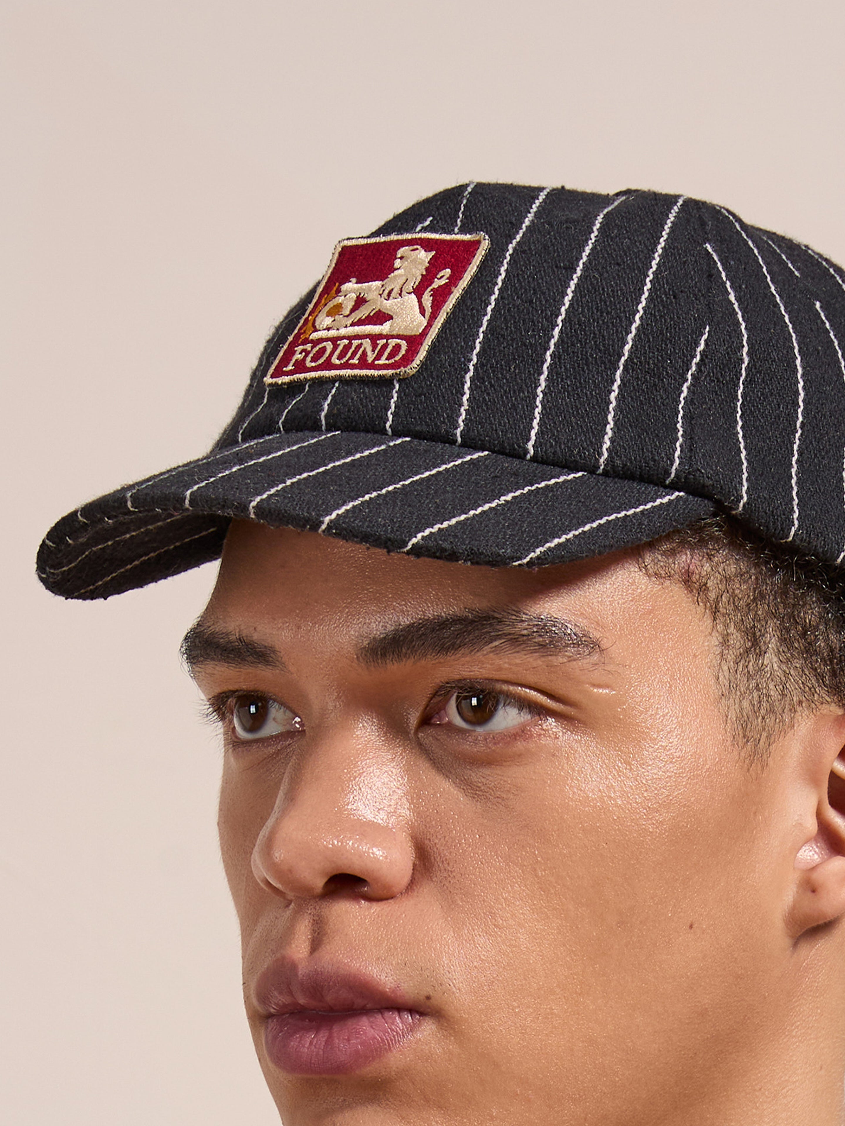 A person wearing the FOUND Pinstripe Lion Cap in washed black, featuring a red and beige patch with a seated figure.