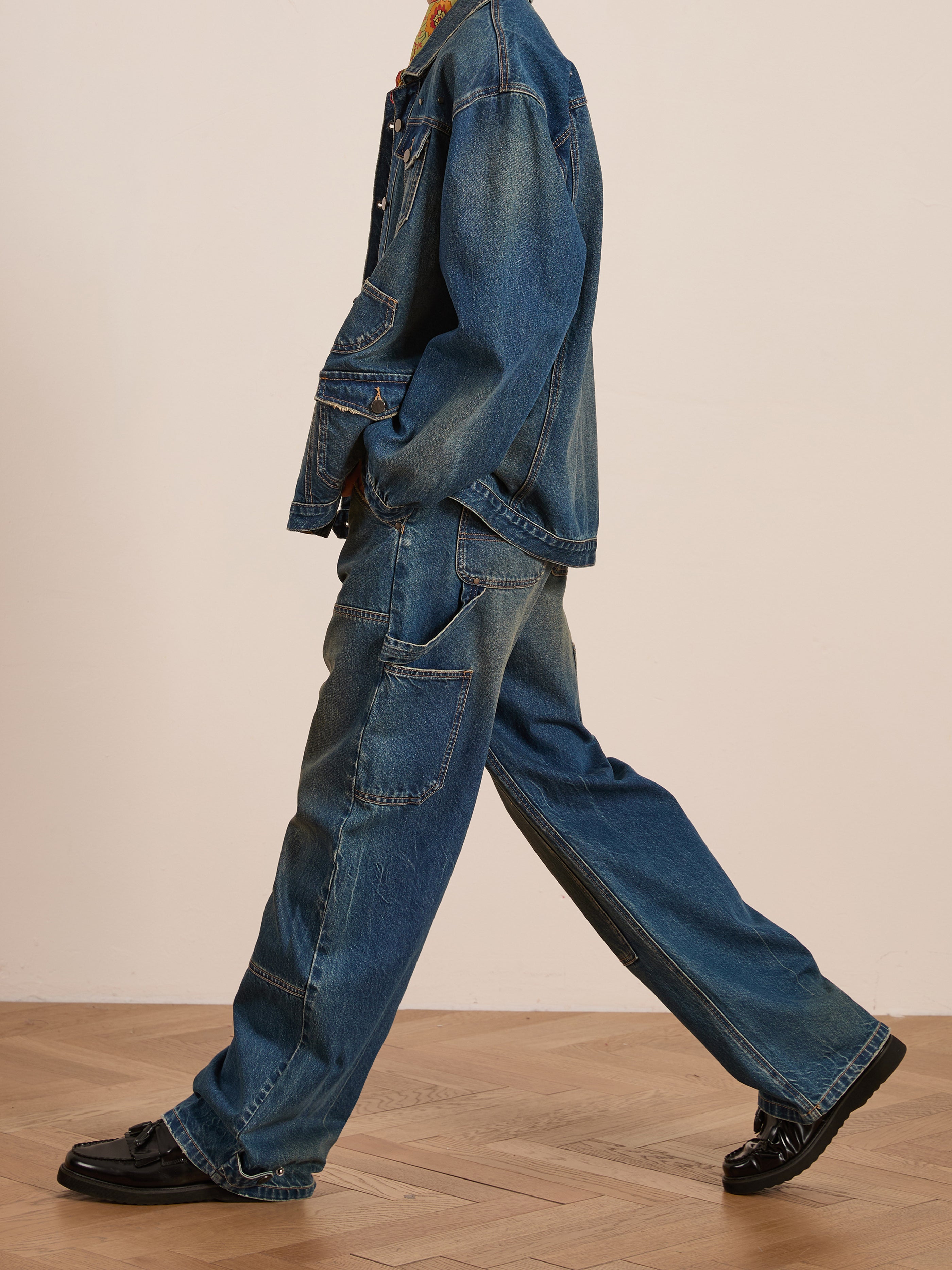 A person in a double denim outfit, wearing the Denim Carpenter Jacket by FOUND, and black shoes, walks on a wooden floor.