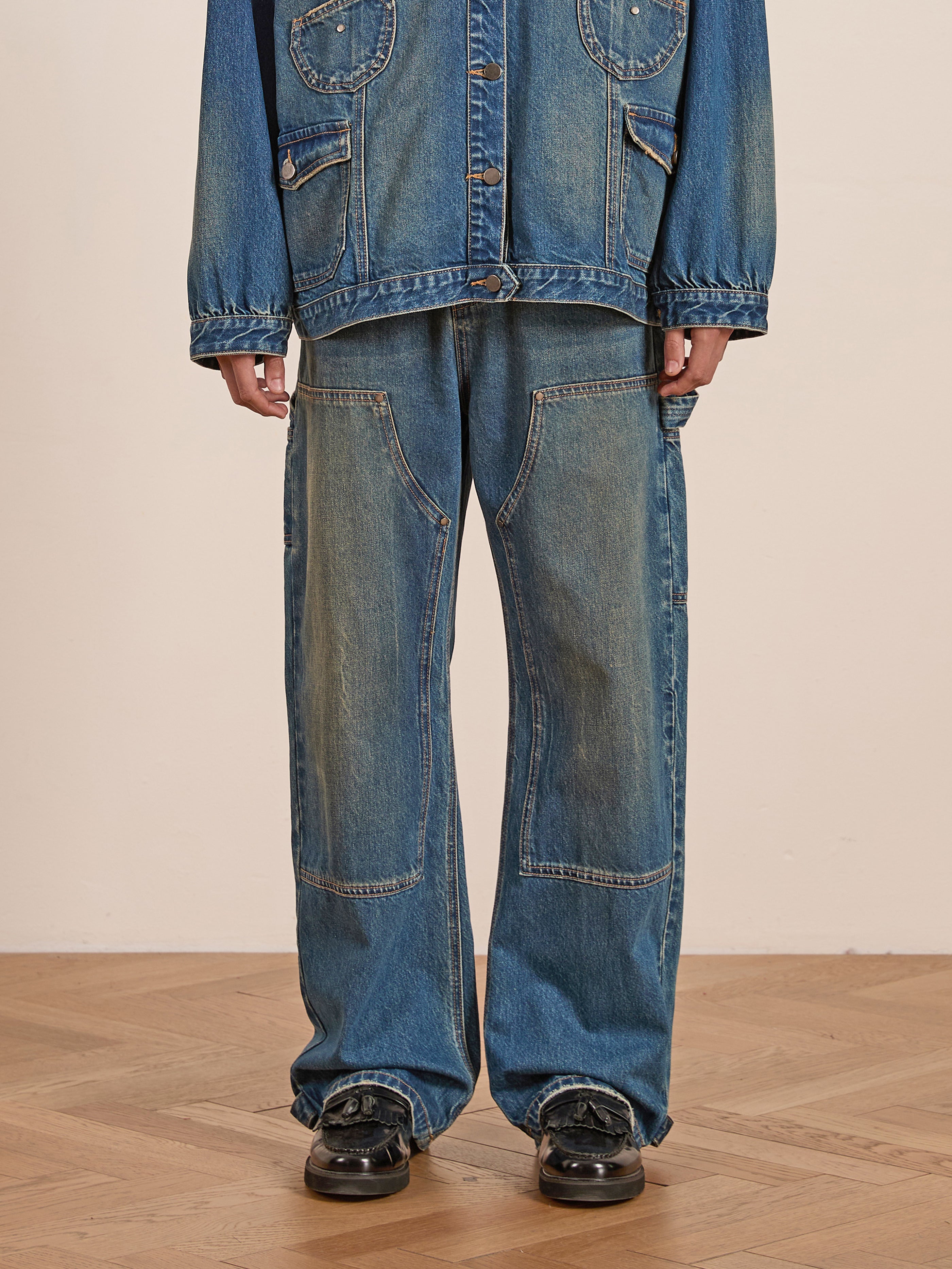 A person wearing an oversized denim jacket with utility pockets and FOUND's Denim Carpenter Jeans stands on a wooden floor, completing the outfit with black shoes.