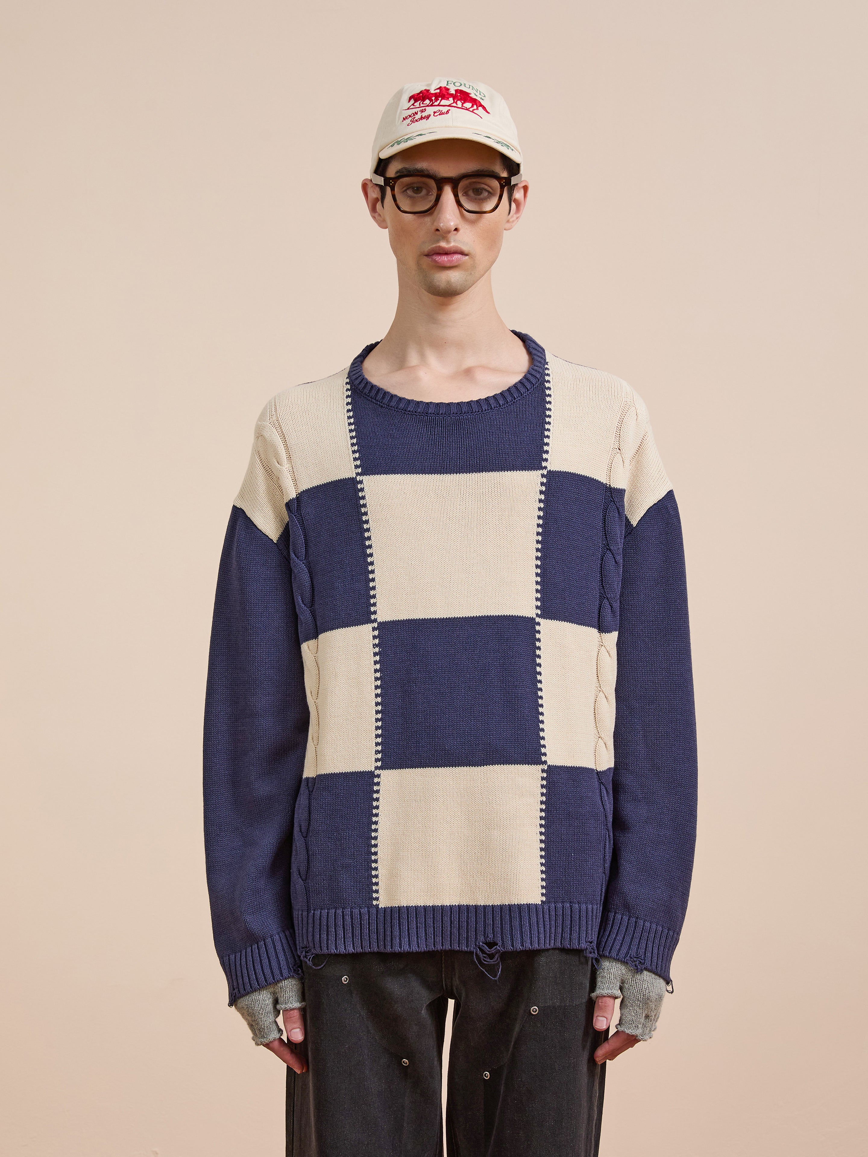A person in glasses, a white cap, fingerless gloves, and dark pants models the Found Checkered Fisherman Sweater against a plain beige background.