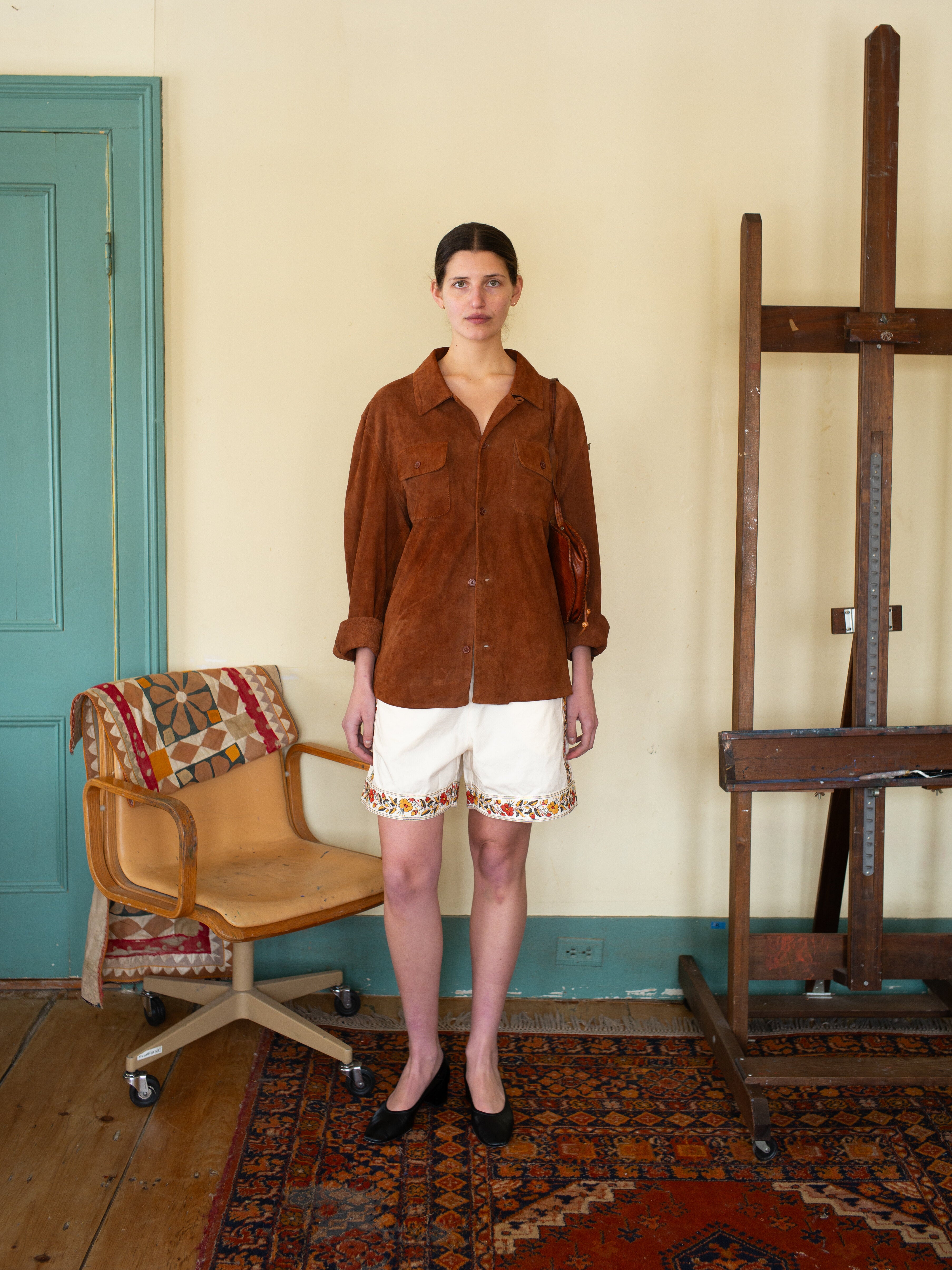 Indoors, a woman stands by a wooden easel wearing FOUND’s brown Suede Leather Camp Shirt with a unisex relaxed fit and white embroidered shorts, alongside a patterned chair and rug.
