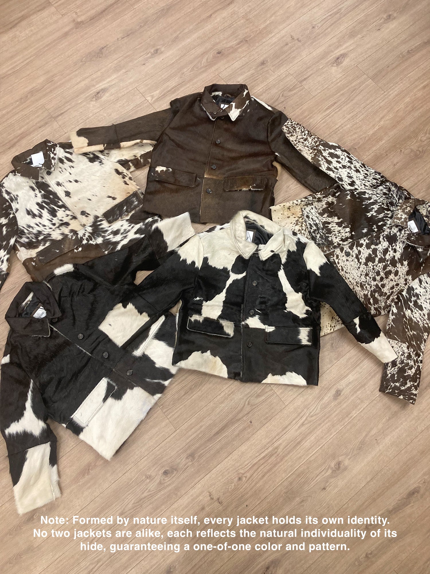 Five FOUND Cowhide Prairie Utility Jackets with unique black, brown, and white patterns are arranged on a wooden floor, each genuine leather piece highlighting its distinct natural design.