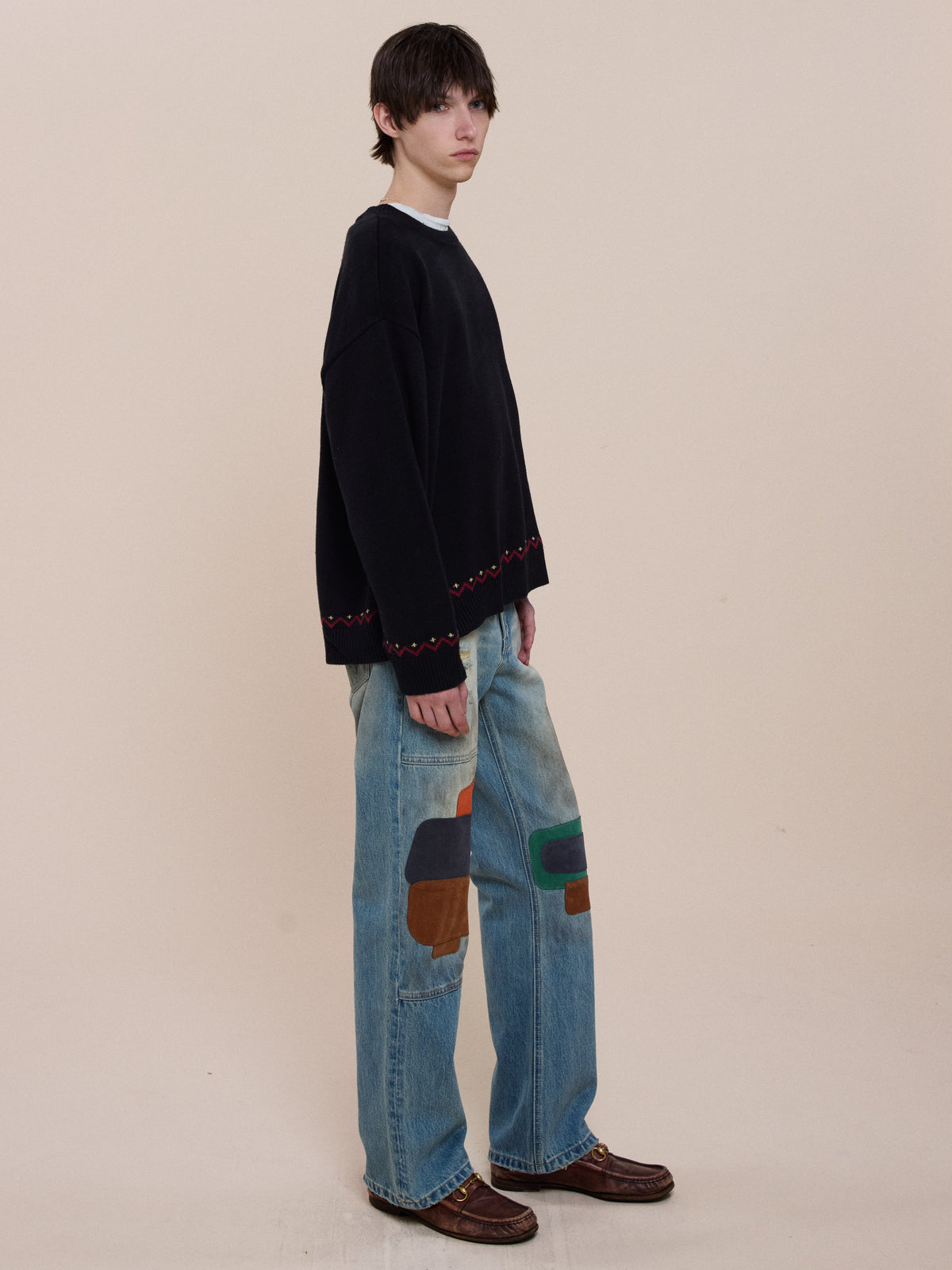 A person stands sideways in a black FOUND Crest Sweater and wide blue jeans with colorful patches, set against a plain beige background.