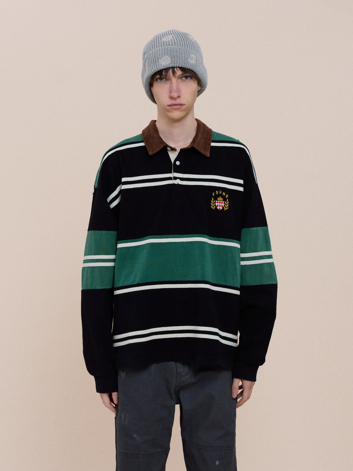 A person wears a gray knit beanie, dark pants, and the FOUND Crestfield Rugby—a unisex shirt with green and white stripes—standing against a beige background.