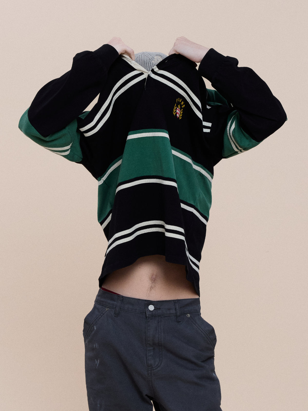 A person pulls a green and black Crestfield Rugby by FOUND over their head, revealing their midriff and grey pants against a plain beige background.