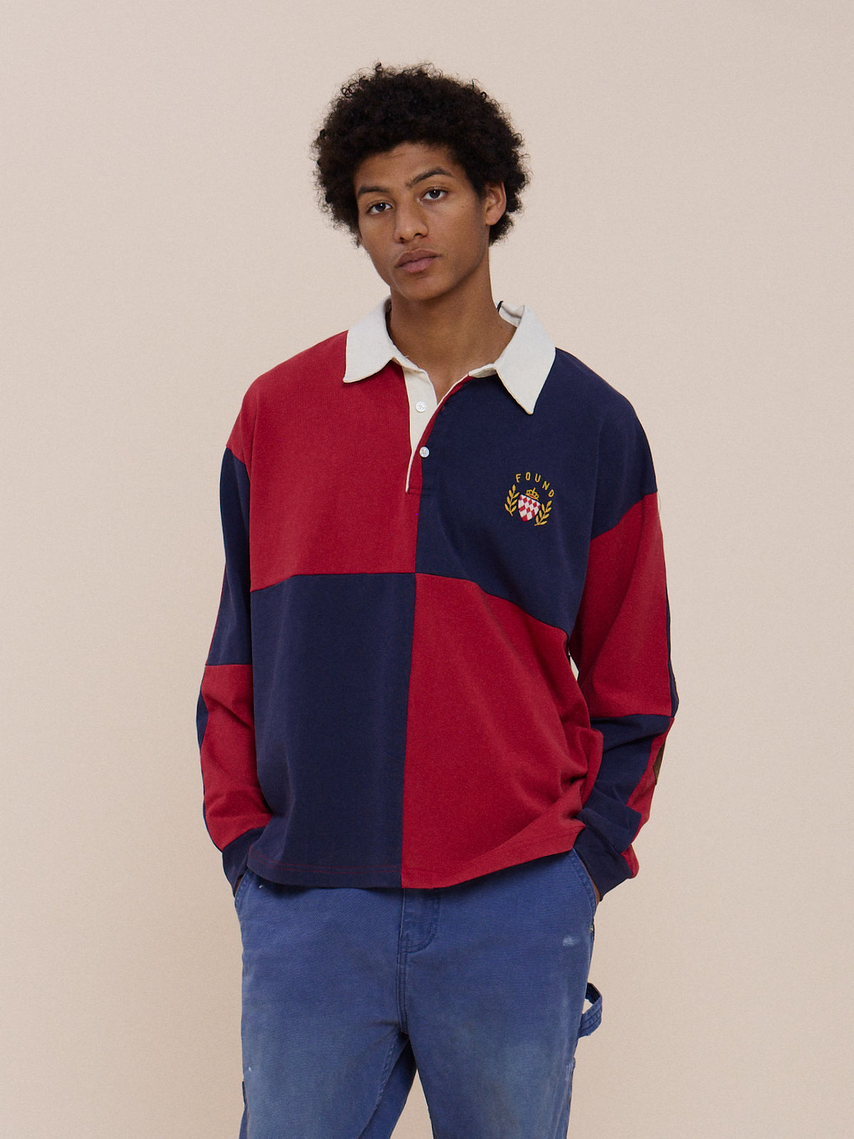 A person wearing the FOUND Crestfield Rugby and blue pants stands against a beige background, highlighting its unisex relaxed fit.