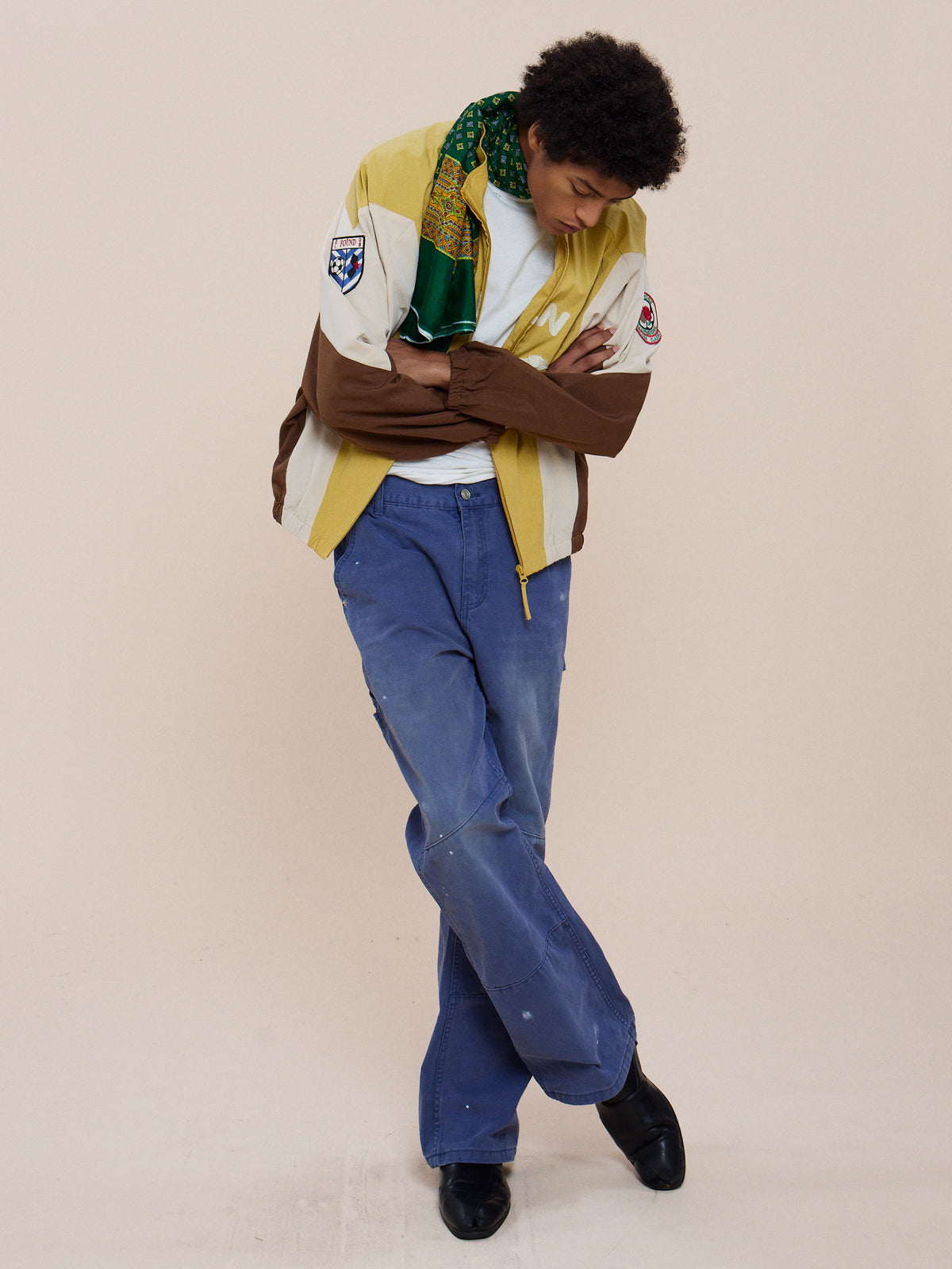 A person in a yellow and brown Crestline Sport Zip Jacket by FOUND, green scarf, white turtleneck, blue jeans, and black shoes poses against a plain beige background.