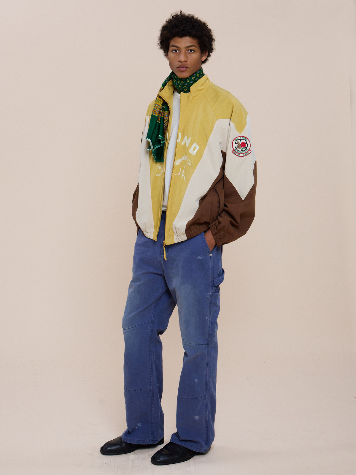 Person stands against a plain background, wearing a yellow and brown Crestline Sport Zip Jacket by FOUND, blue cargo pants, black shoes, and a green patterned scarf.