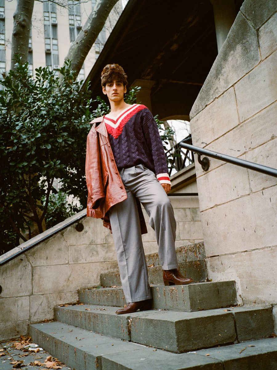 Wearing a FOUND V-Neck Cricket Sweater in navy and red, a person walks down stone steps with gray pants and brown shoes, holding a brown jacket. Trees and a building add elegance to the background.