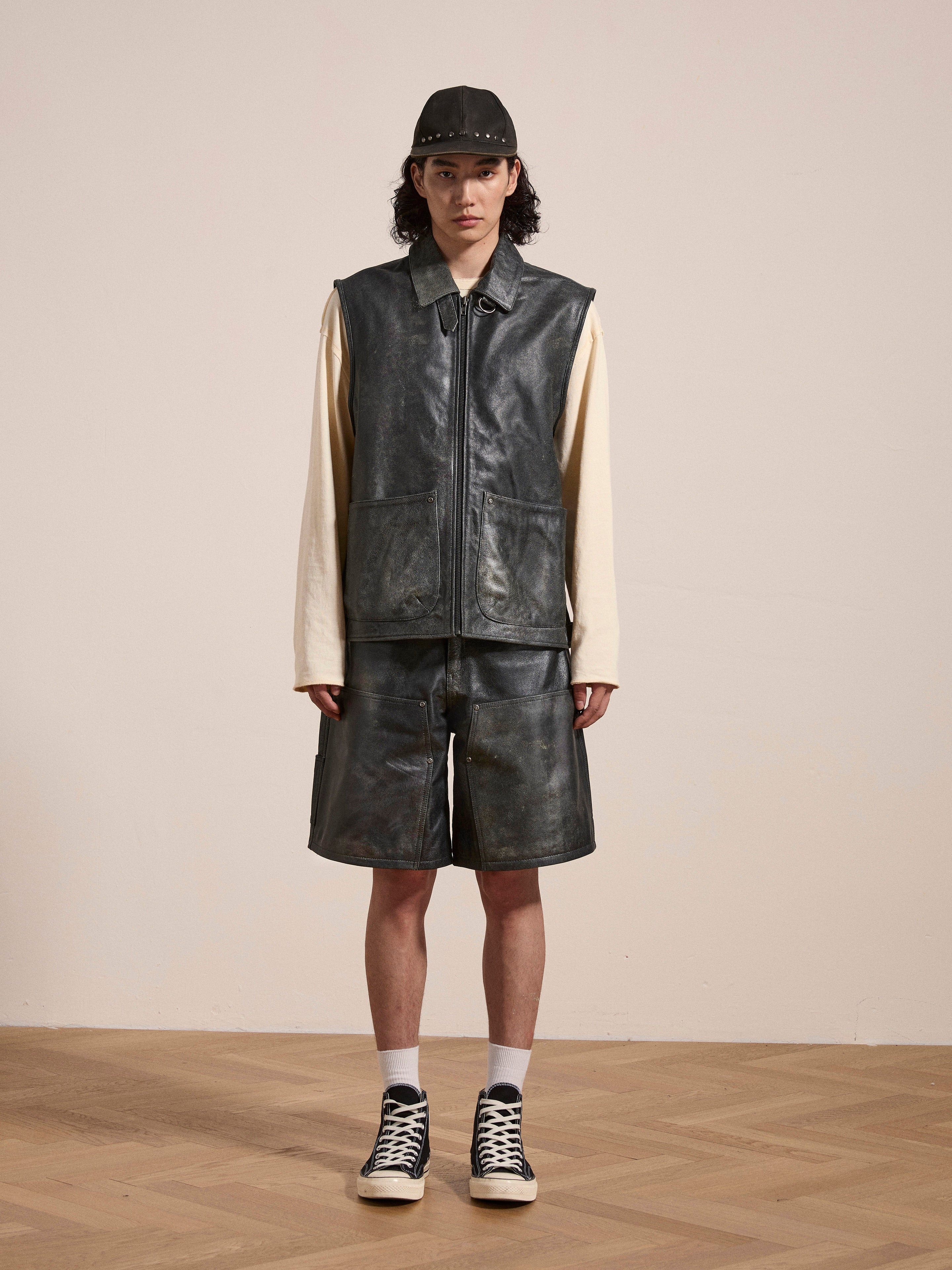 A person stands indoors wearing a black cap, a sleeveless leather vest over a long-sleeve shirt, Found Distressed Genuine Leather Carpenter Shorts, white socks, and black high-top sneakers. The background is plain and minimal.