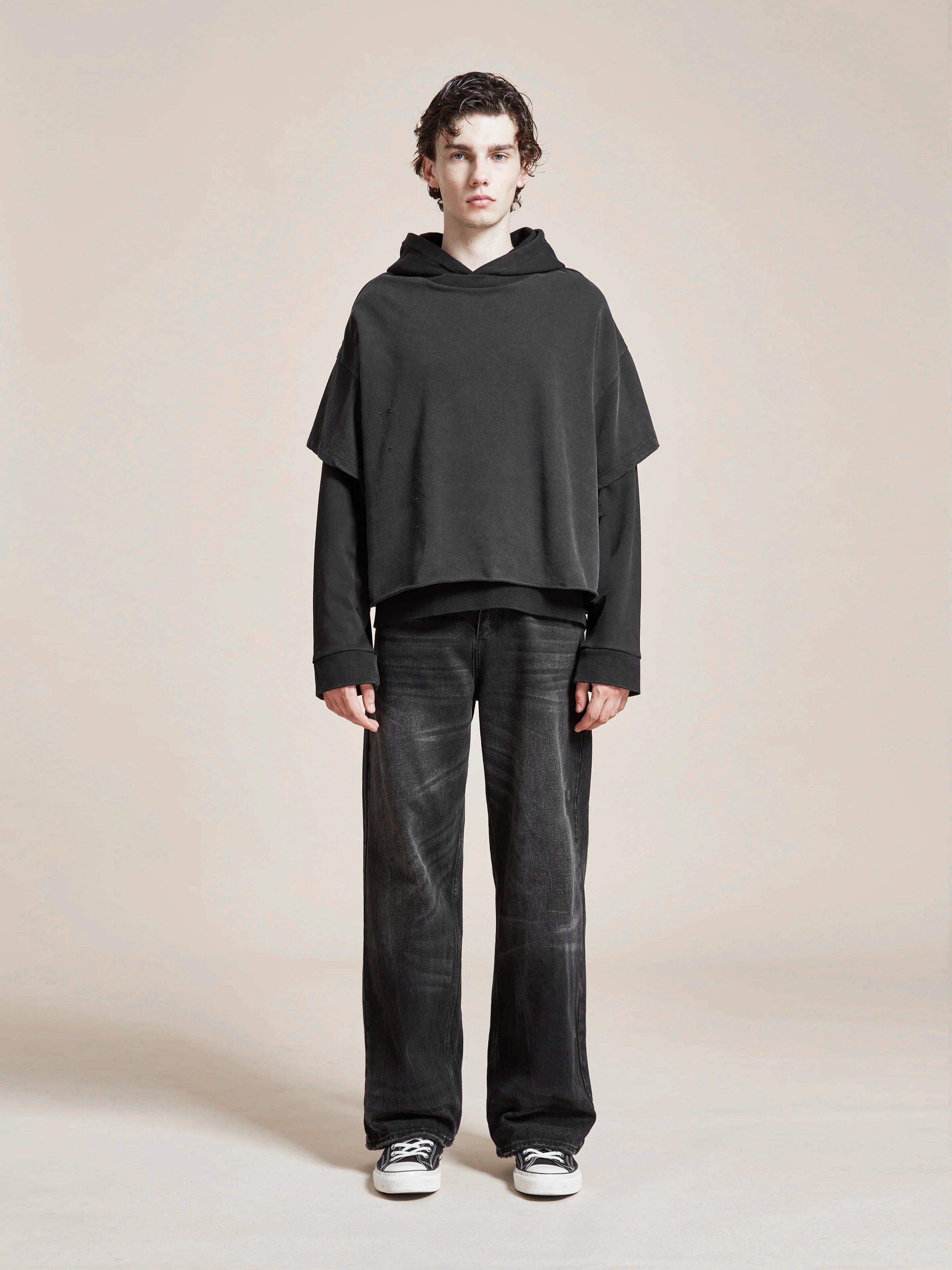 Young man in a black oversized Found Double Layer Hoodie and gray jeans standing against a plain beige background.