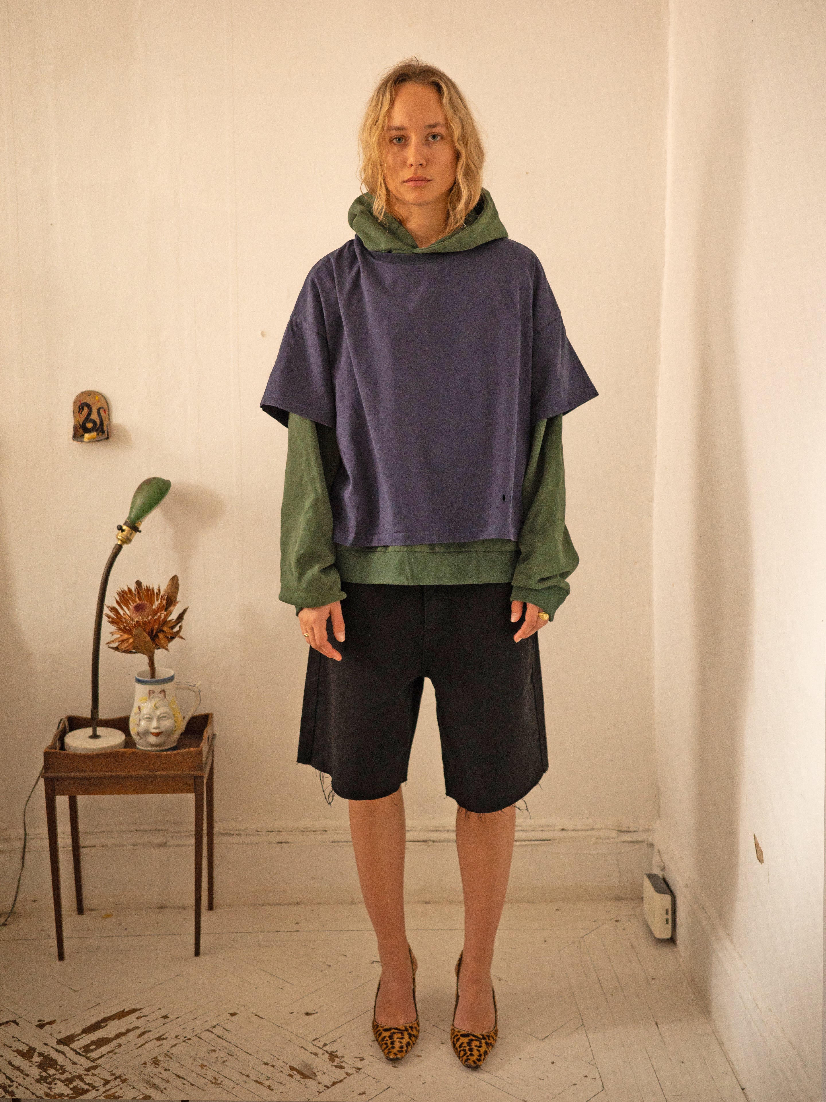 In a room, someone wears the Found Double Layer Hoodie with the green underlayer visible beneath a blue short-sleeve top. They pair it with black shorts and leopard-print heels. A small table with a lamp to the left completes this vintage scene.