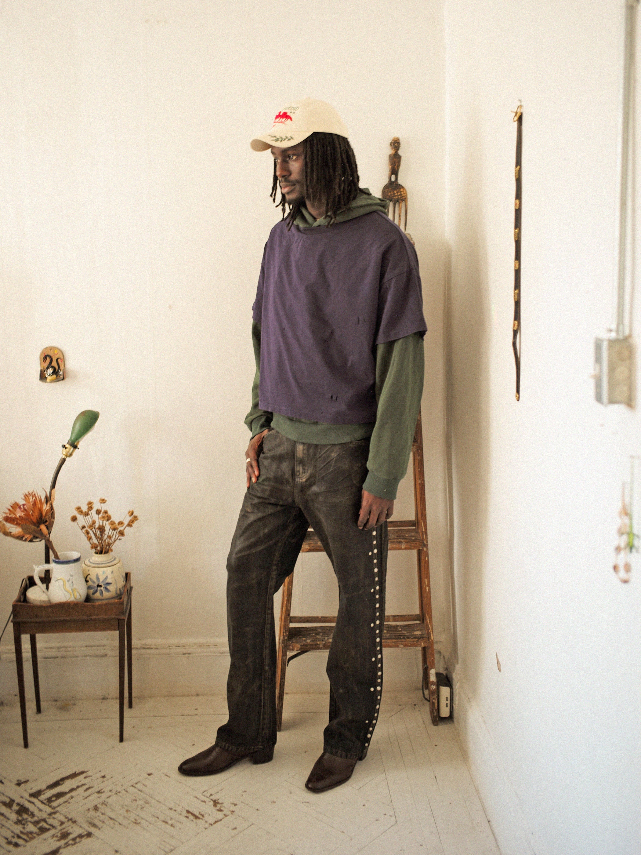 A man in a vintage beige cap, purple shirt over a Found Double Layer Hoodie in green, and studded jeans stands in a room with light walls. To the left is a small table adorned with flowers.