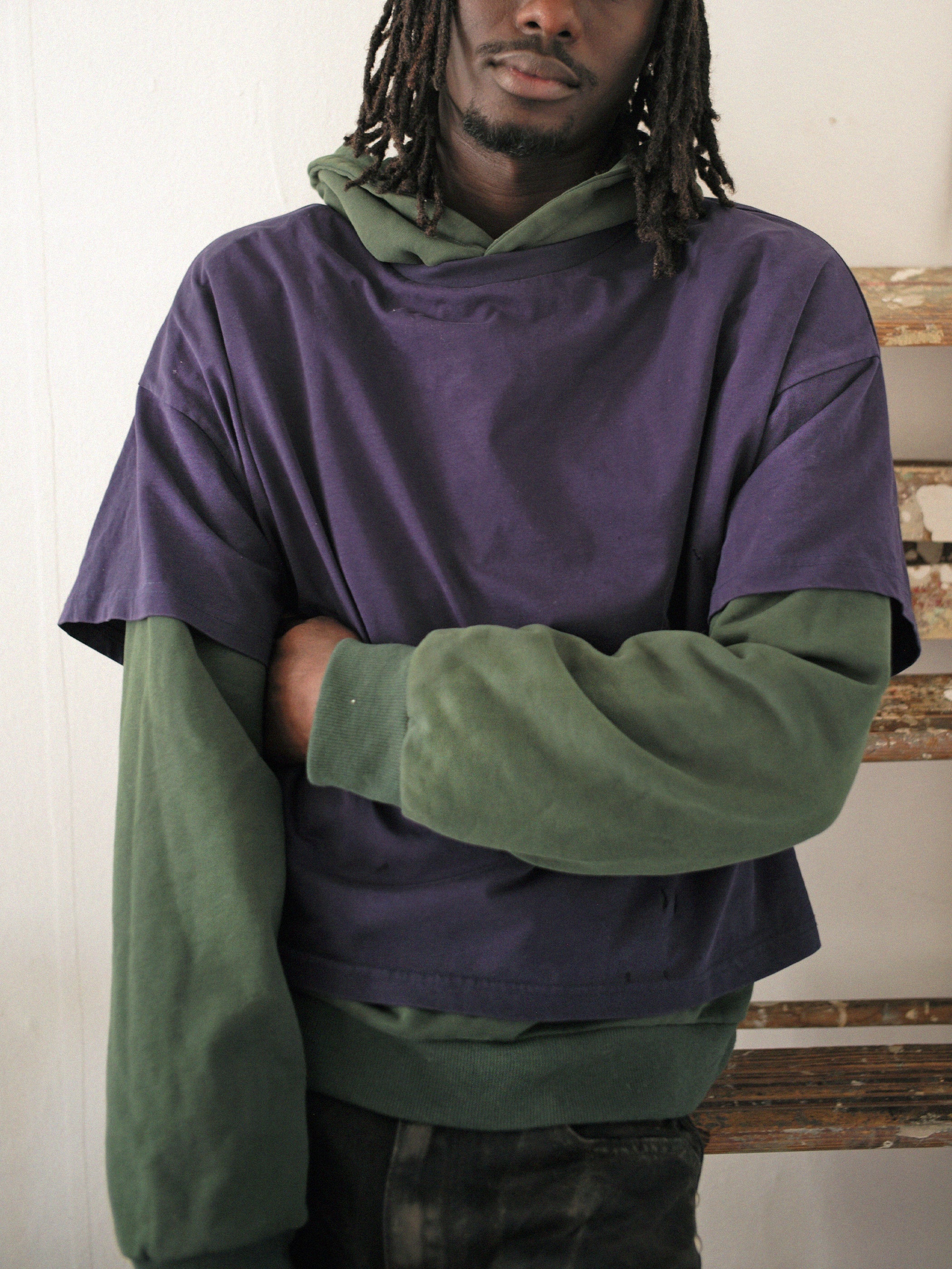 A person in a purple short-sleeved shirt over Found's vintage-look green Double Layer Hoodie stands indoors with one arm crossed.