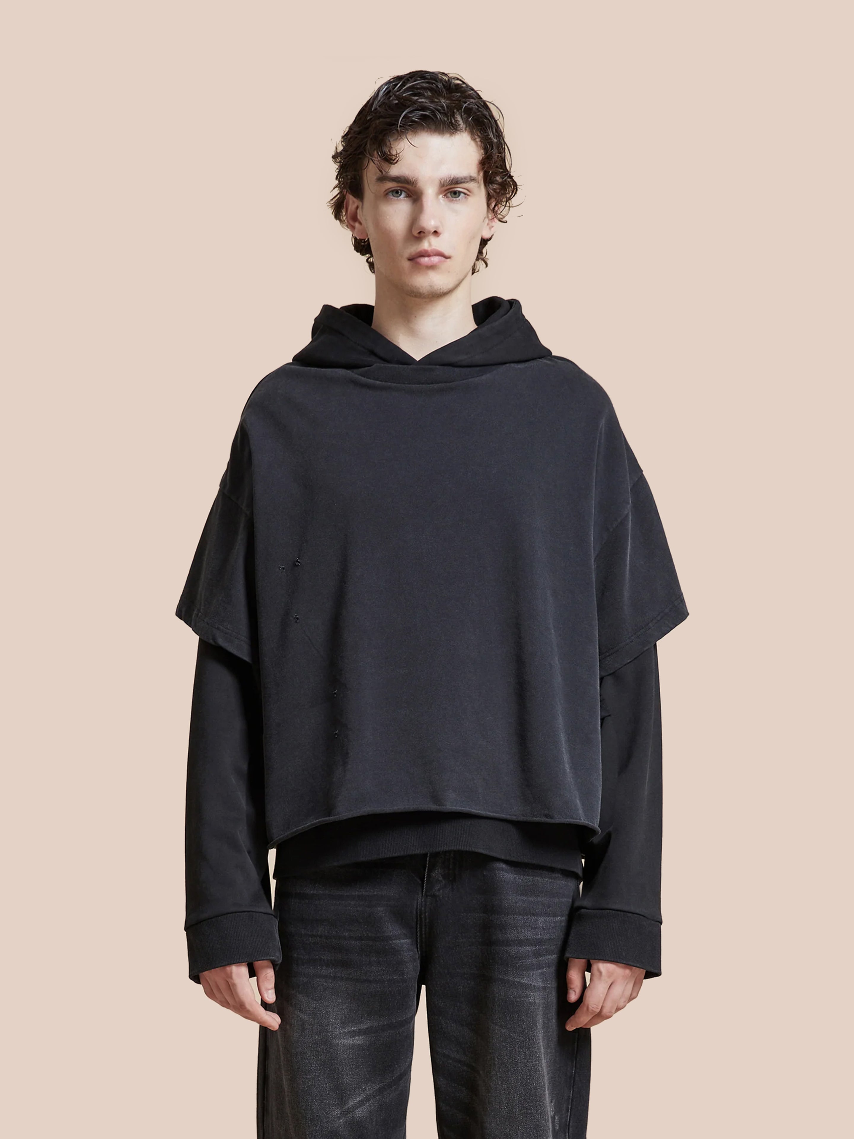 A young person with short, wavy brown hair models the Found Double Layer Hoodie in french terry cotton and black jeans, standing against a plain beige background.