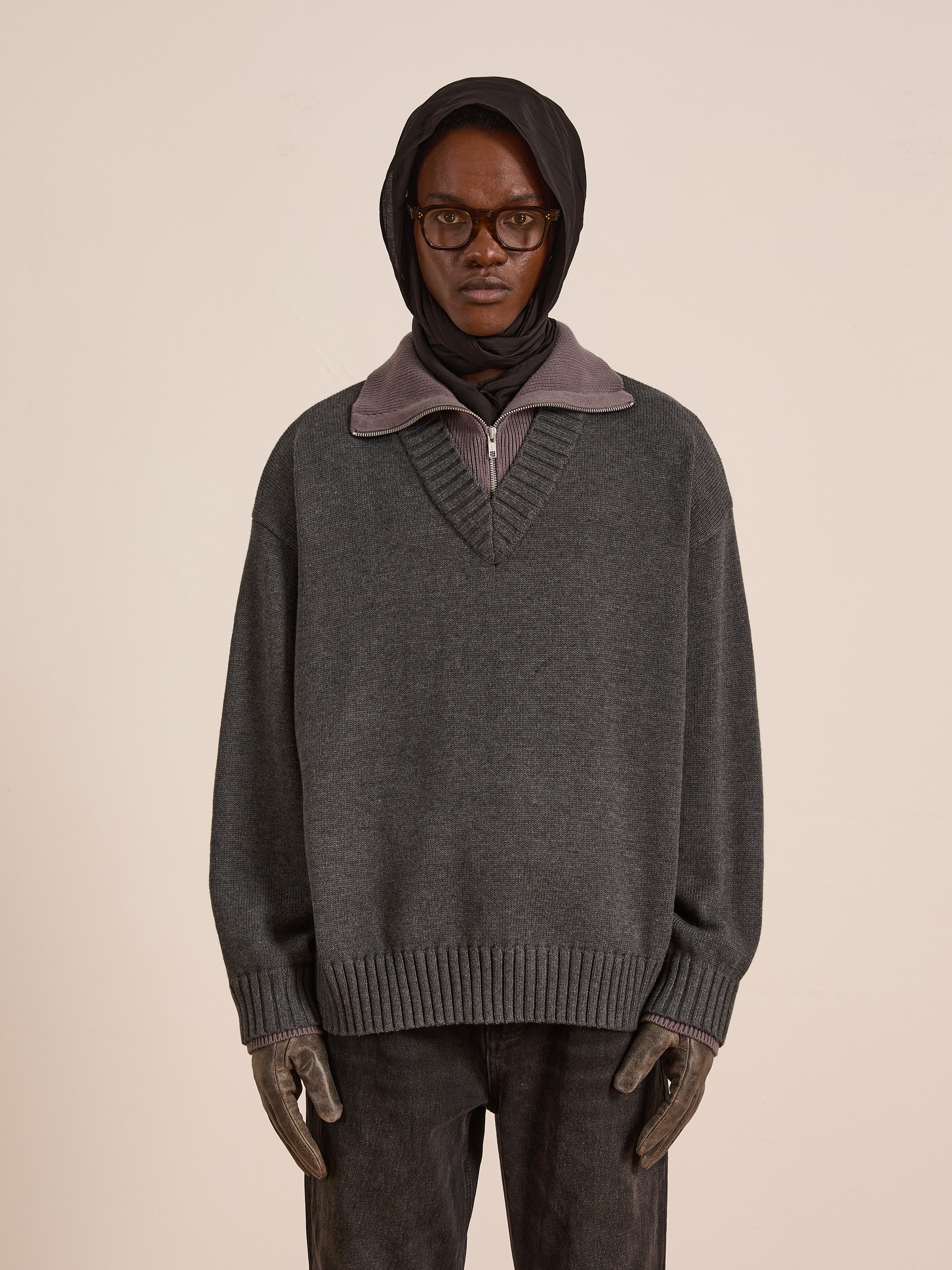 A person in glasses and a headscarf wears the FOUND Double Layer Sweater, an oversized grey wool blend, with gloves and dark pants against a plain beige background—ideal for cold-weather layering.