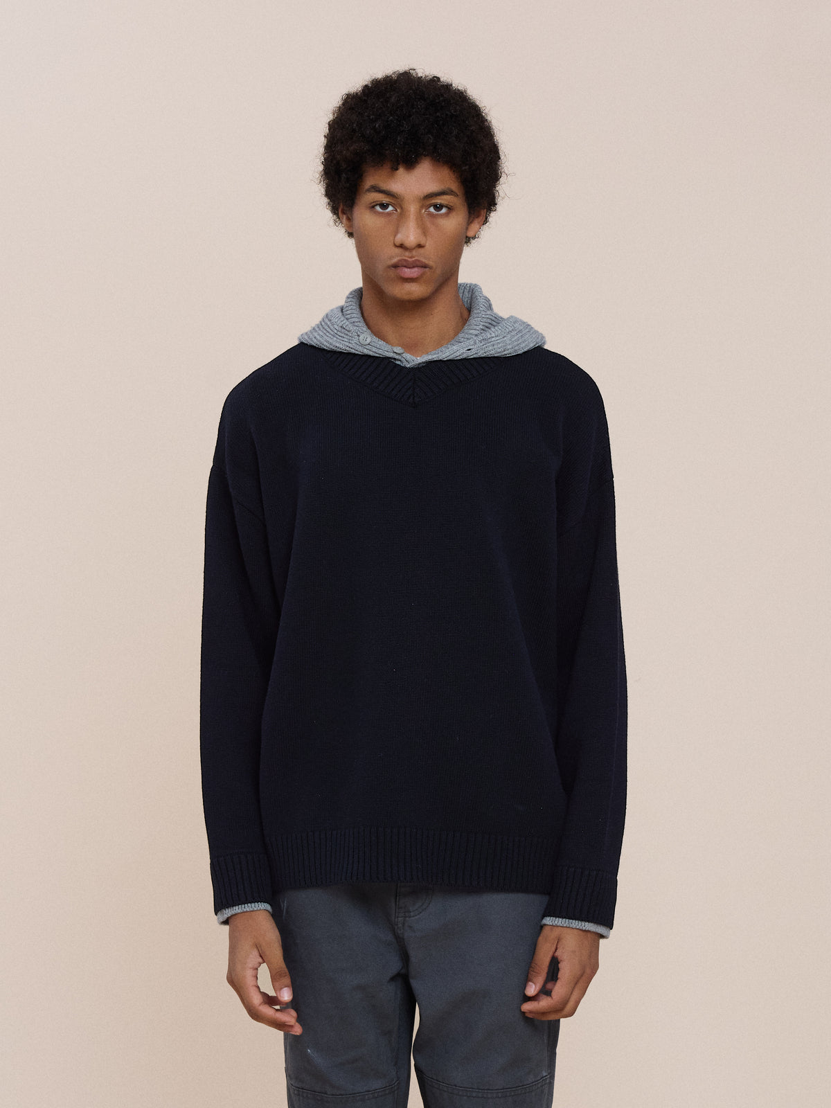 A person with short curly hair wears the FOUND Double Layer V Neck Knit Sweater over a gray hoodie and dark pants, standing against a plain beige background.