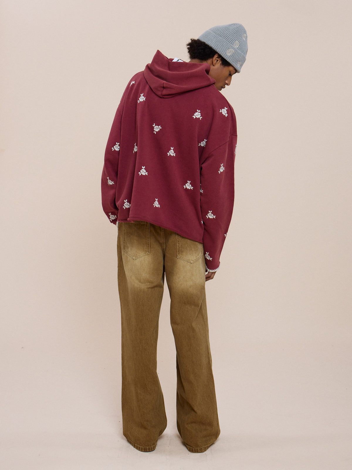 Person stands with back to camera, wearing FOUND Embroidered Motif Hoodie in maroon, beige wide-leg pants, and a light blue knit beanie against a plain background.