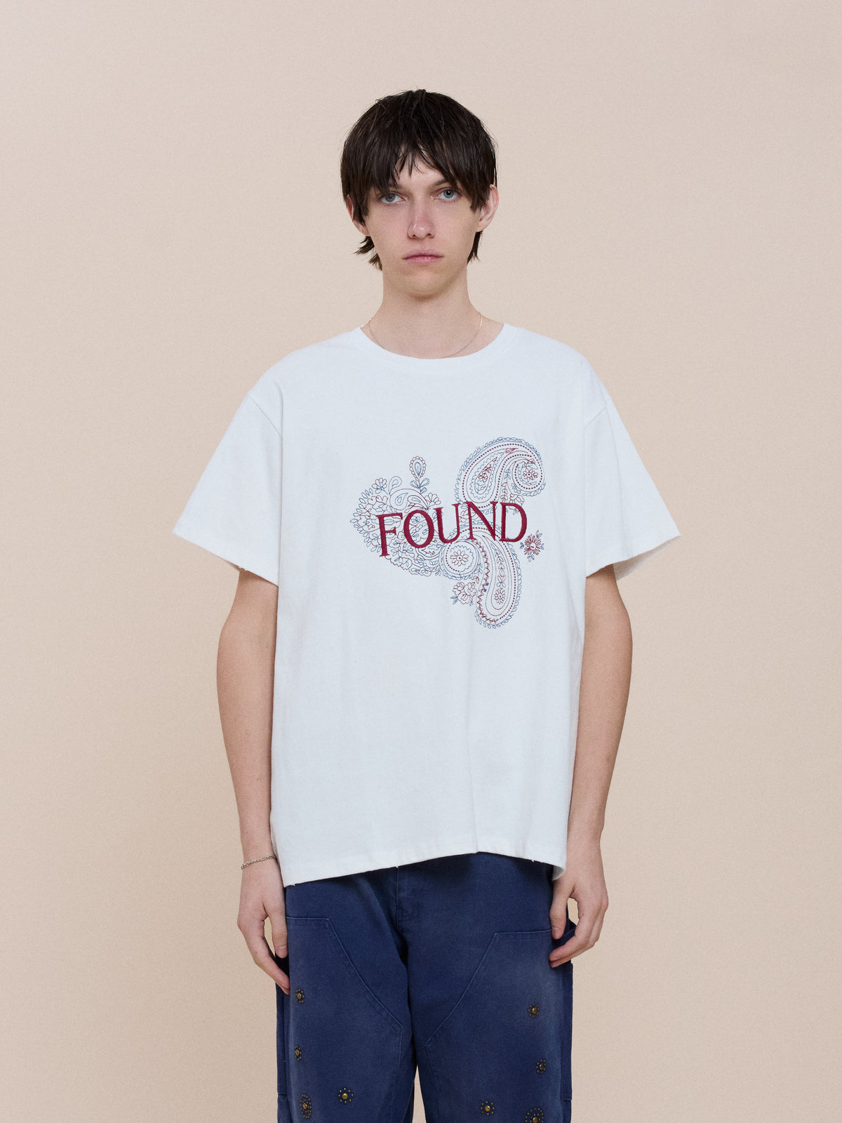 Person with short, dark hair wears the Embroidered Paisley Tee by FOUND, featuring a bandana-inspired paisley print, paired with blue pants against a beige background.