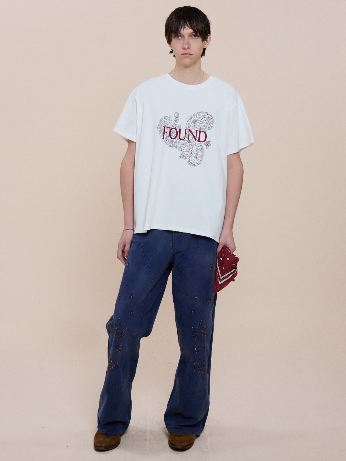 Person stands against a beige background, wearing the FOUND Embroidered Paisley Tee, blue embellished pants, brown shoes, and holding a red item.