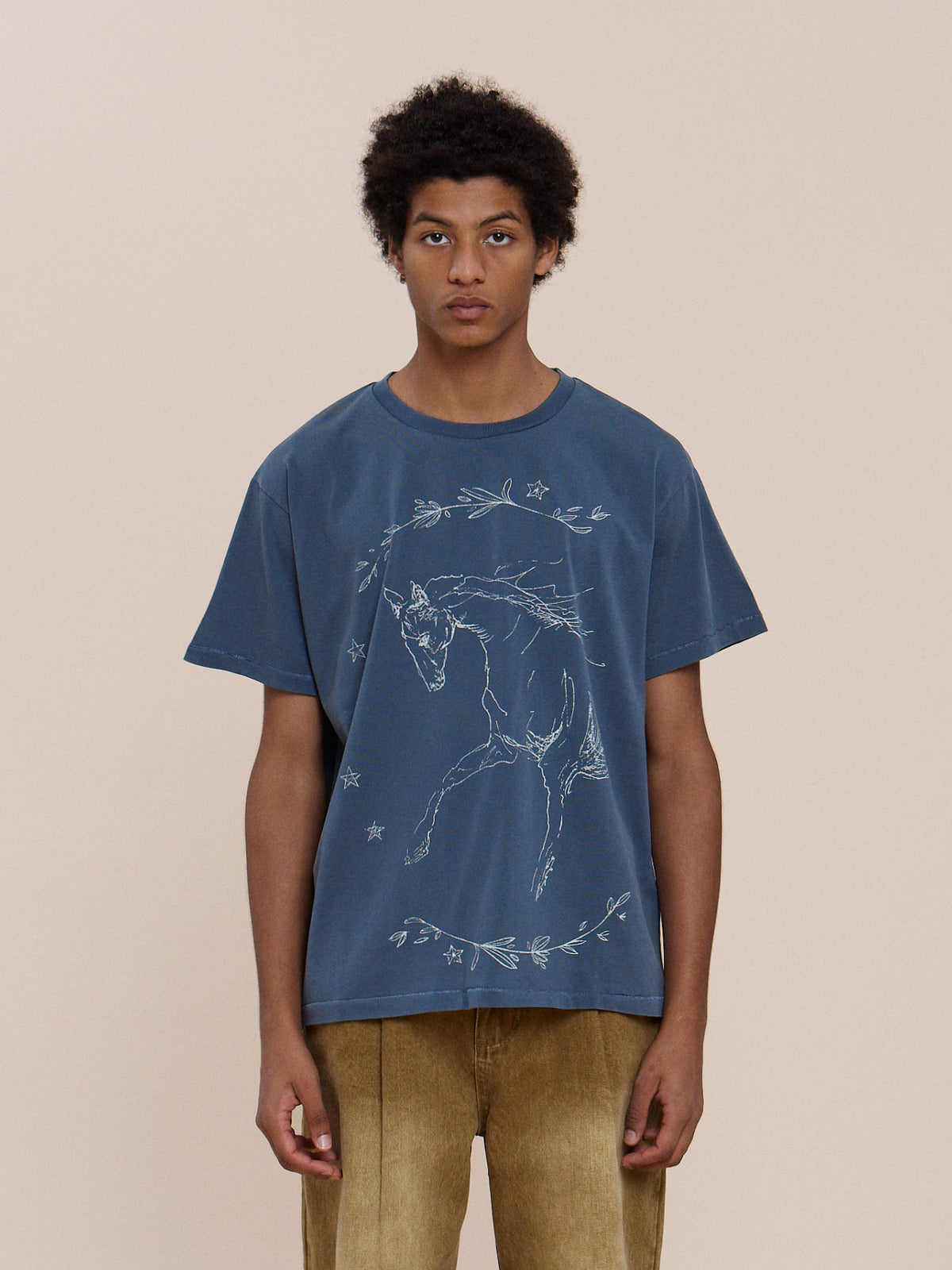 A person stands facing forward in the FOUND Faded Horse Trot Tee, a unisex relaxed fit blue shirt with a white horse and floral print, paired with tan pants.
