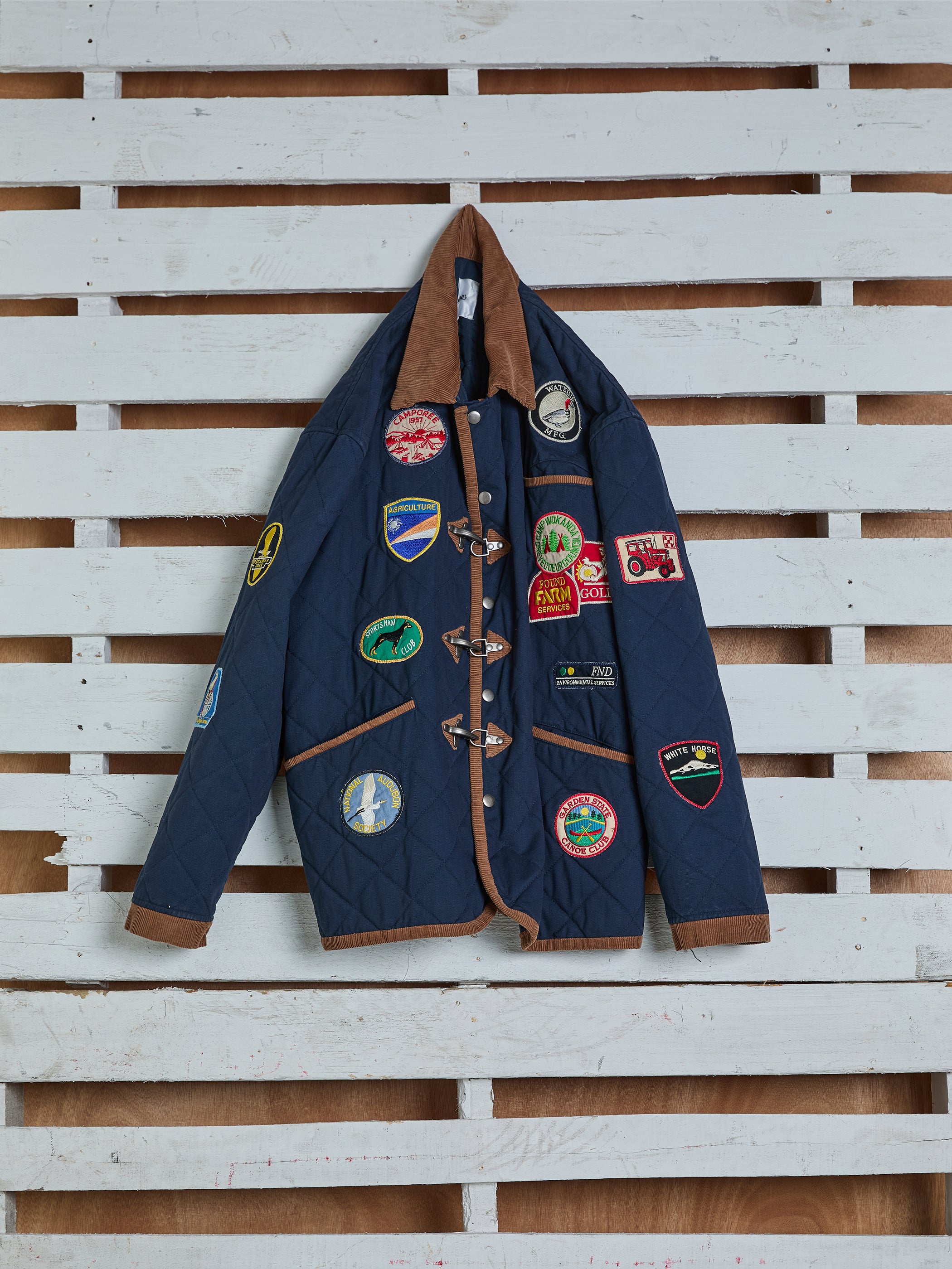 A blue Farmstead Quilt Patch Jacket by FOUND, featuring colorful patches and brown trim for a vintage hunting vibe, hangs against a white wooden wall. This jacket boasts a relaxed unisex fit.