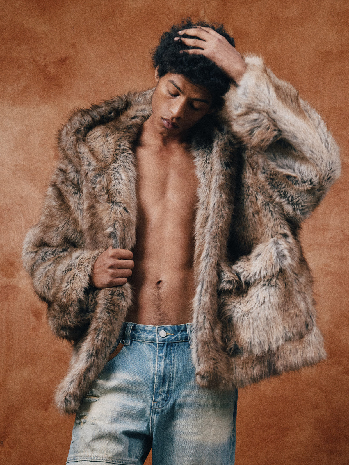 A shirtless person in vintage FOUND Faux Fur Hoodie and jeans stands against a brown background, touching their hair and holding the hoodie open.