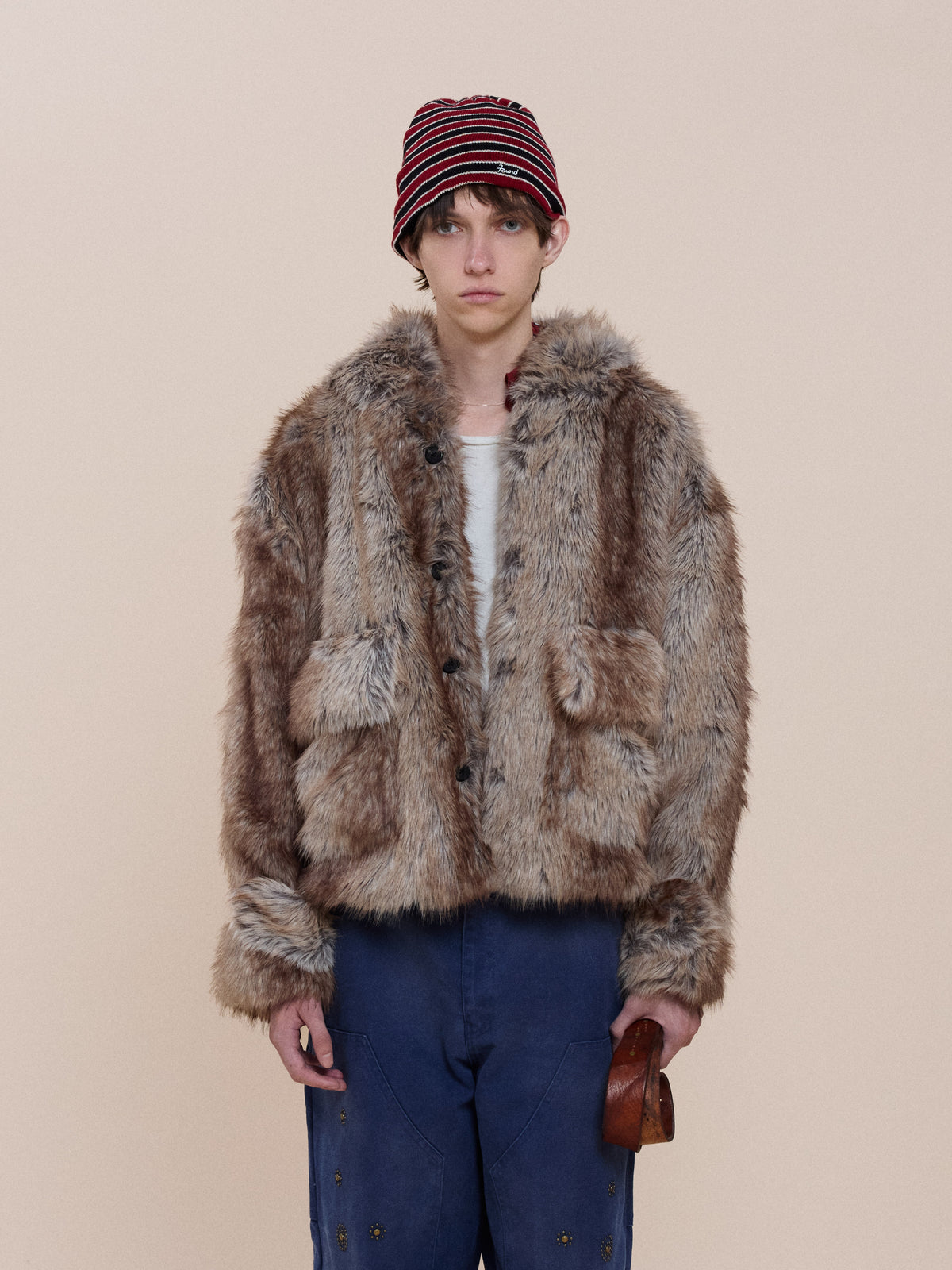 Person wearing the FOUND Faux Fur Hoodie, white shirt, blue pants, and a red-striped beanie stands against a plain beige background.