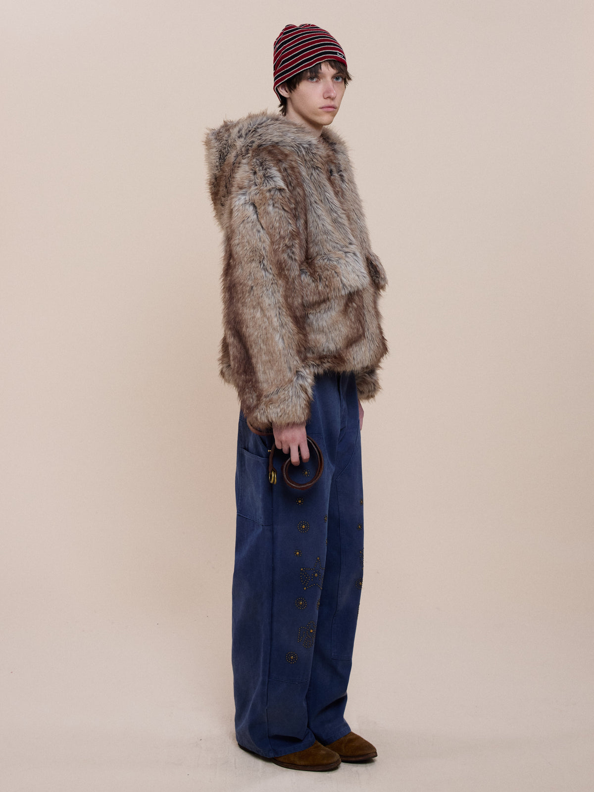 A person in the FOUND Faux Fur Hoodie, wide-leg blue grommet pants, brown shoes, and a red-striped beanie holds a round object against a beige background.