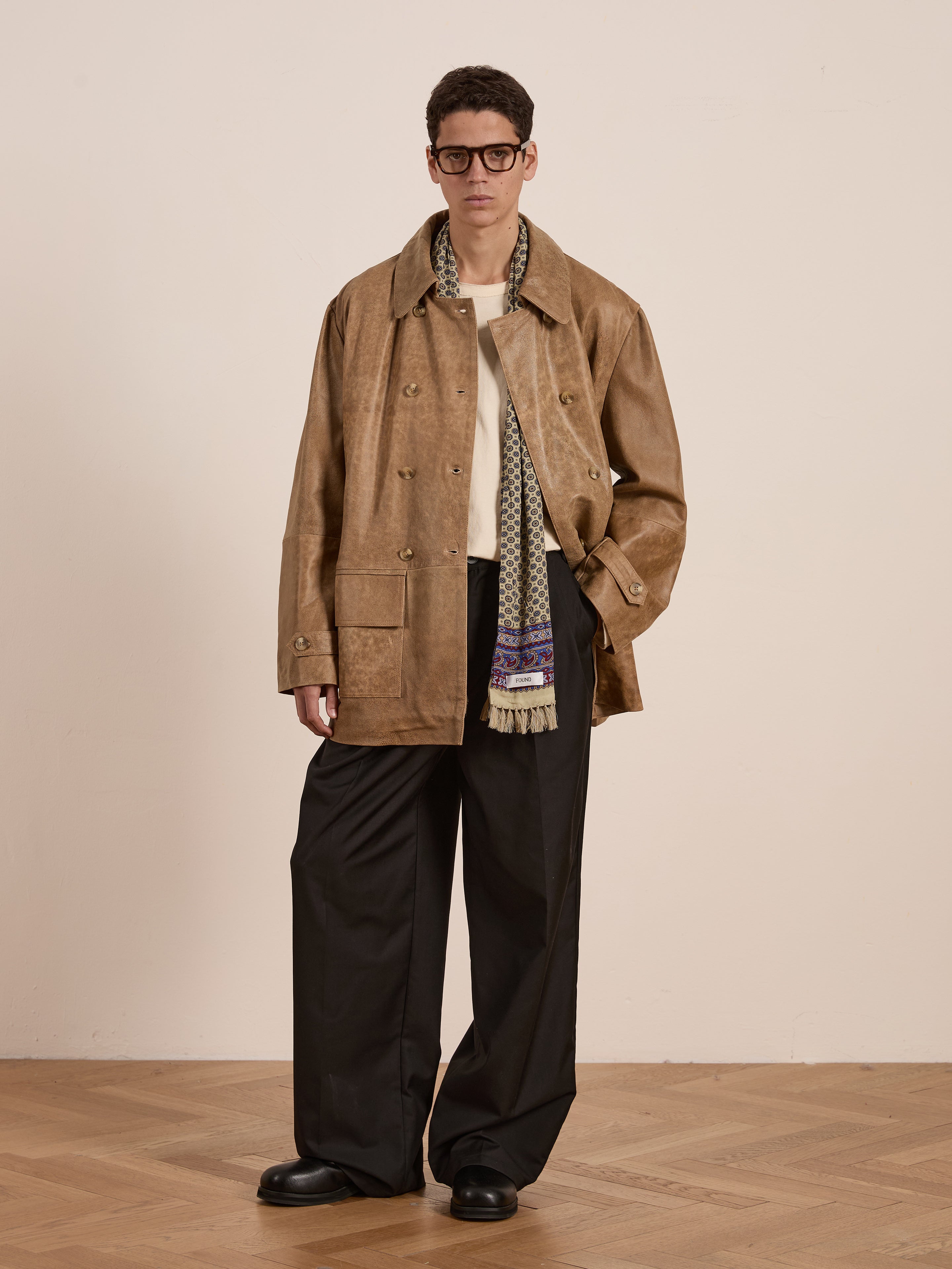 A person stands on a wooden floor against a plain wall, wearing the ethically produced Fawn Double Breasted Leather Jacket by FOUND. The look is finished with a patterned scarf, white shirt, black wide-leg pants, and black shoes.