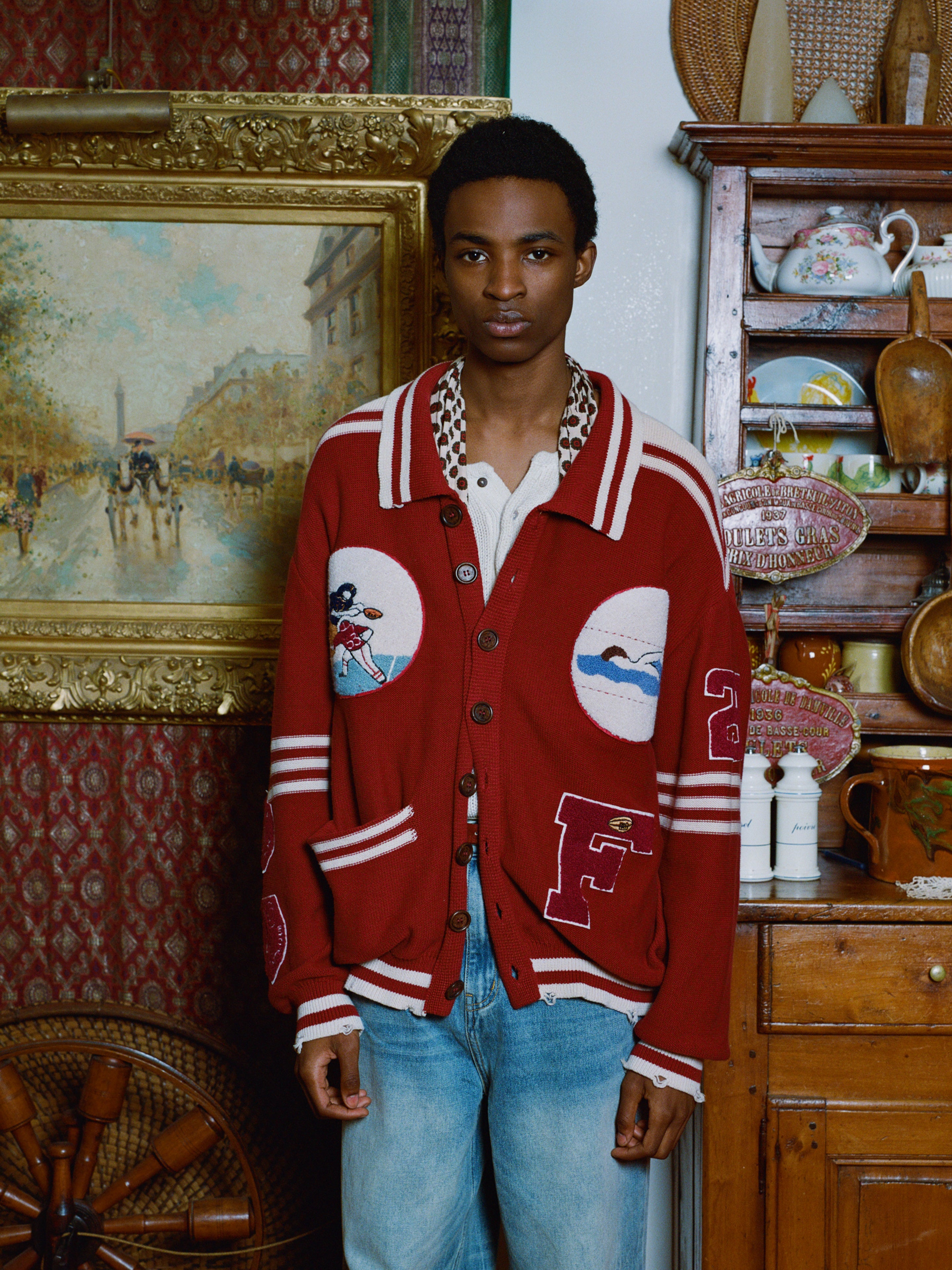 A person in a Fin Varsity Patch Collared Cardigan by Found and jeans stands indoors near a wooden cabinet and framed artwork, capturing a vintage look.