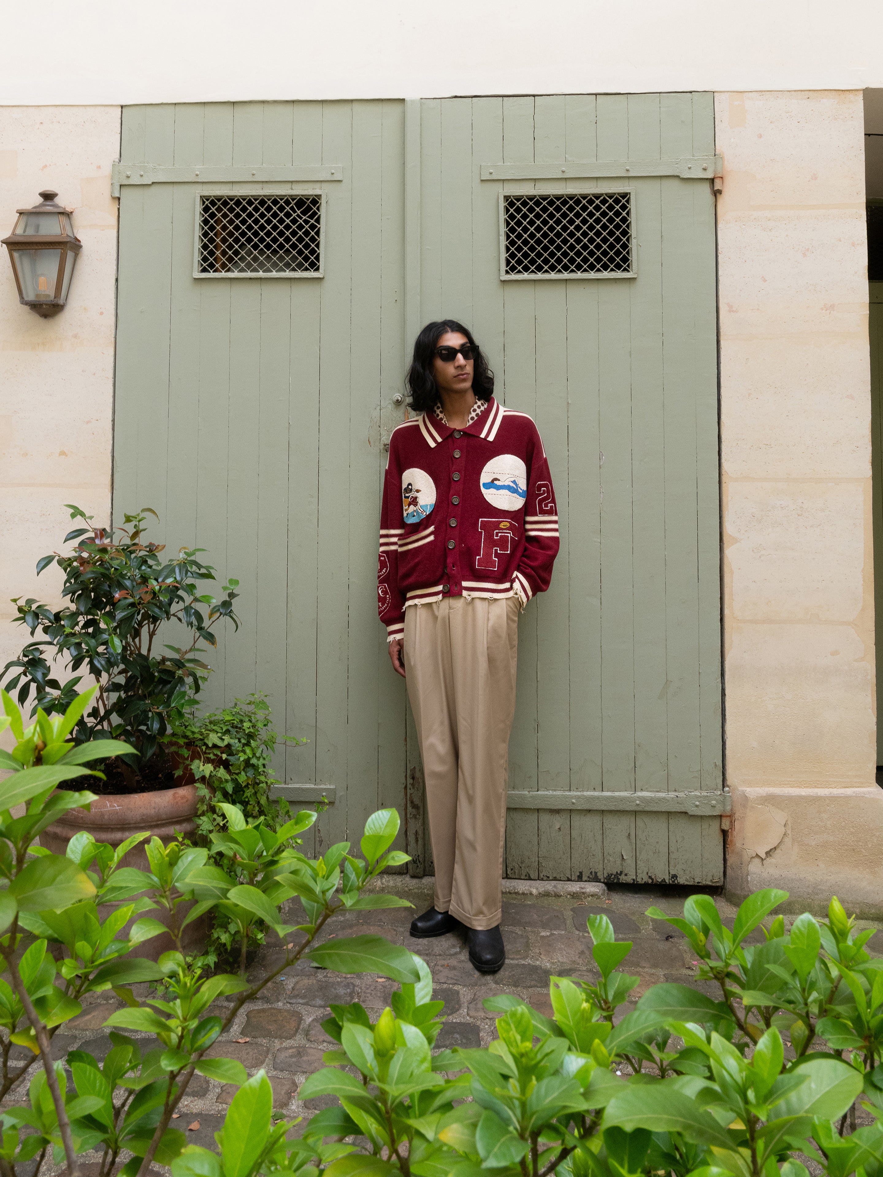 A person in a Found Fin Varsity Patch Collared Cardigan, beige pants, and sunglasses stands against a green door, exuding a vintage vibe among vibrant potted plants.