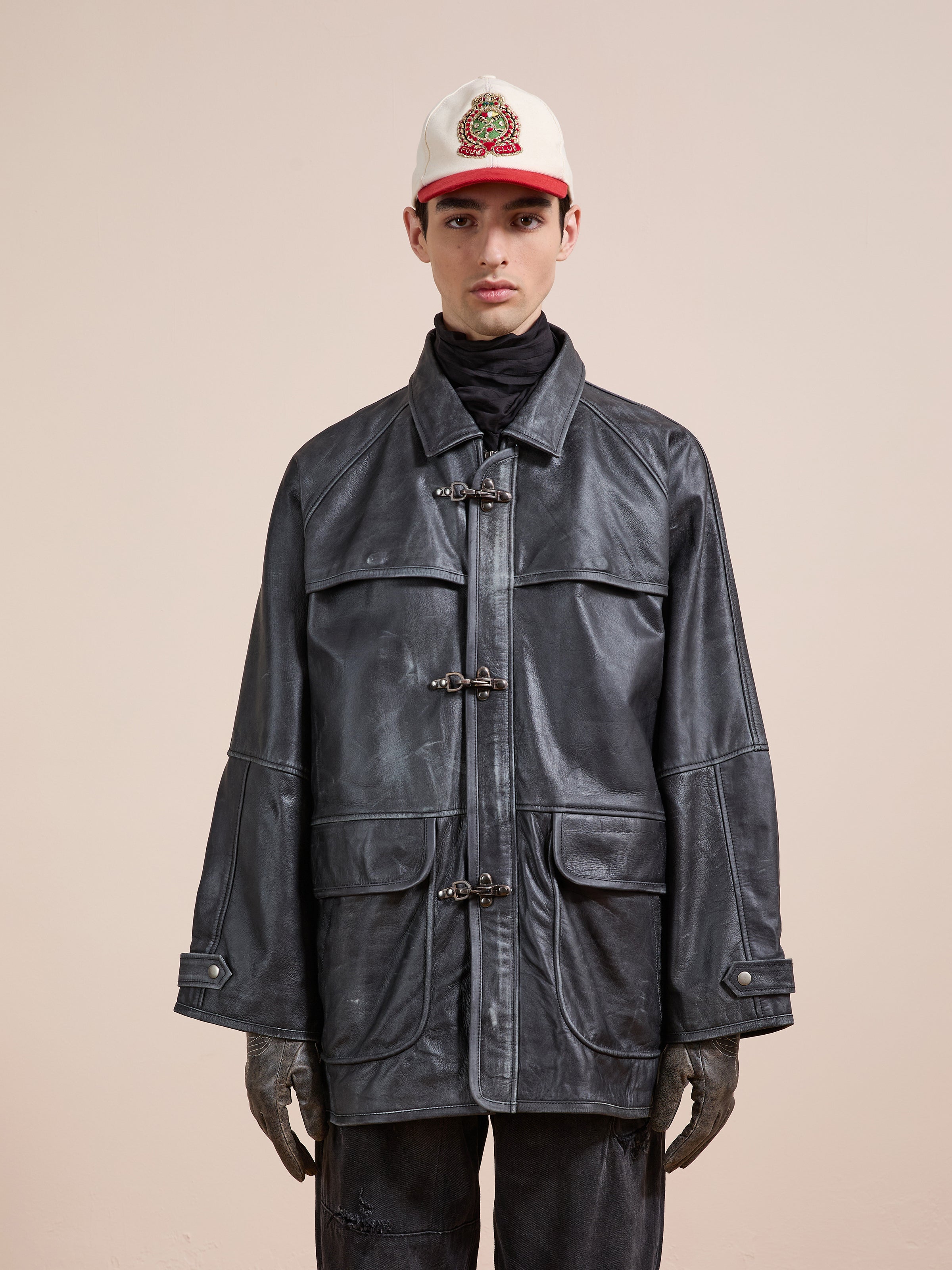 A person in a white cap, black turtleneck, black gloves, dark pants, and the FOUND Distressed Leather Fireman Clasp Jacket (unisex relaxed fit) stands against a plain beige background.