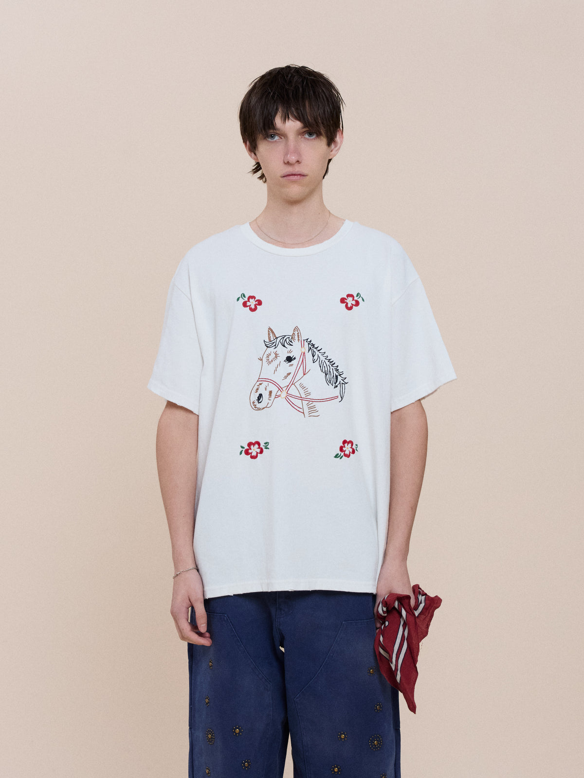 A person stands against a beige background in the FOUND Floral Horse Tee with a vintage folk textiles design, blue pants, and holds a red patterned cloth in their right hand.