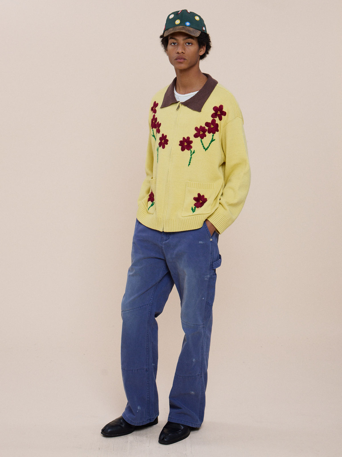 Person wears a green bucket hat, FOUND Floral Sun Knit Sweater, blue jeans, and black shoes against a plain light background.