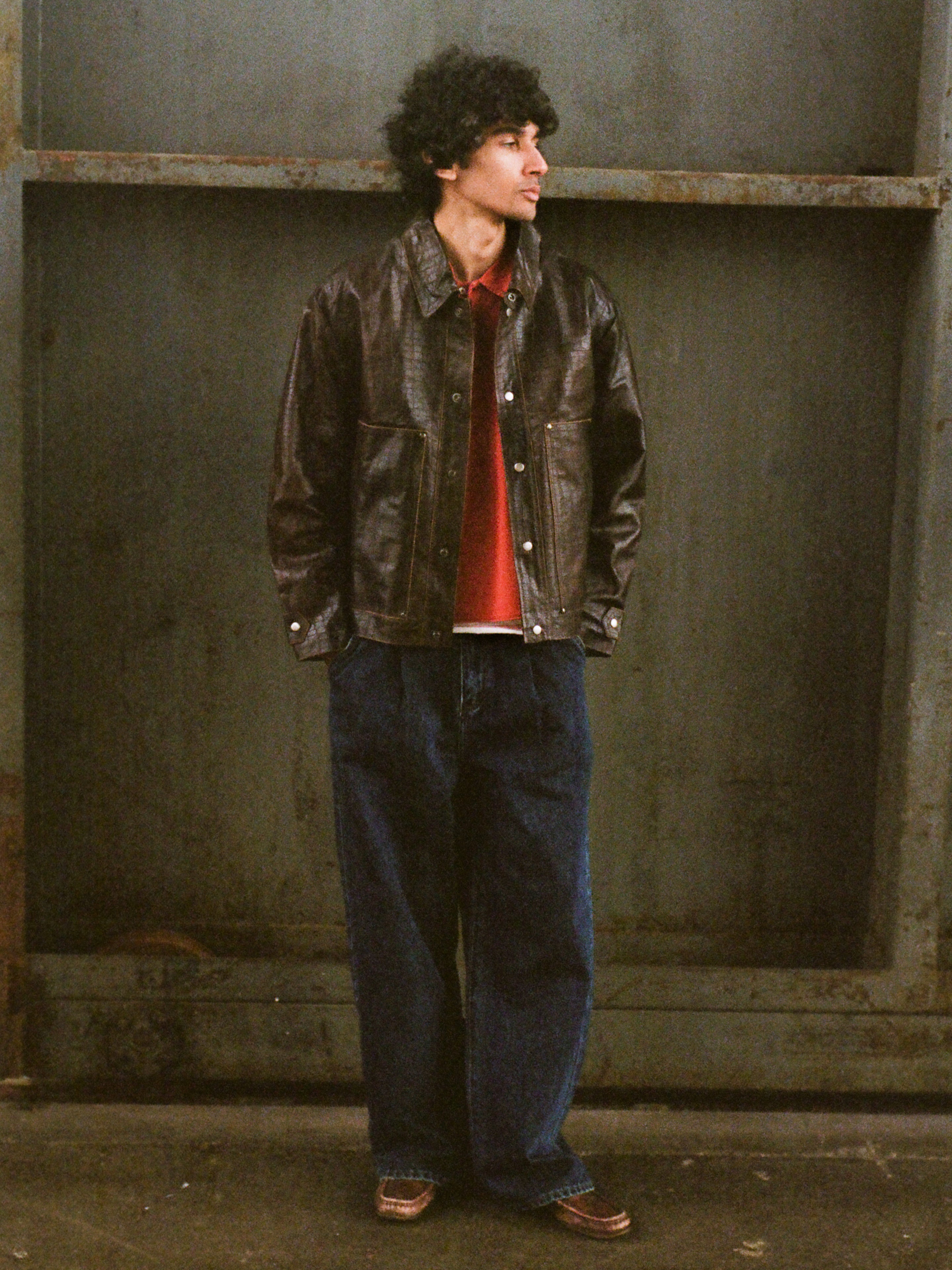 A person with curly hair wears the FOUND Distressed Croc Embossed Leather Work Jacket, a red shirt, wide-leg jeans, and brown shoes while standing in front of a metal wall and looking to the side.