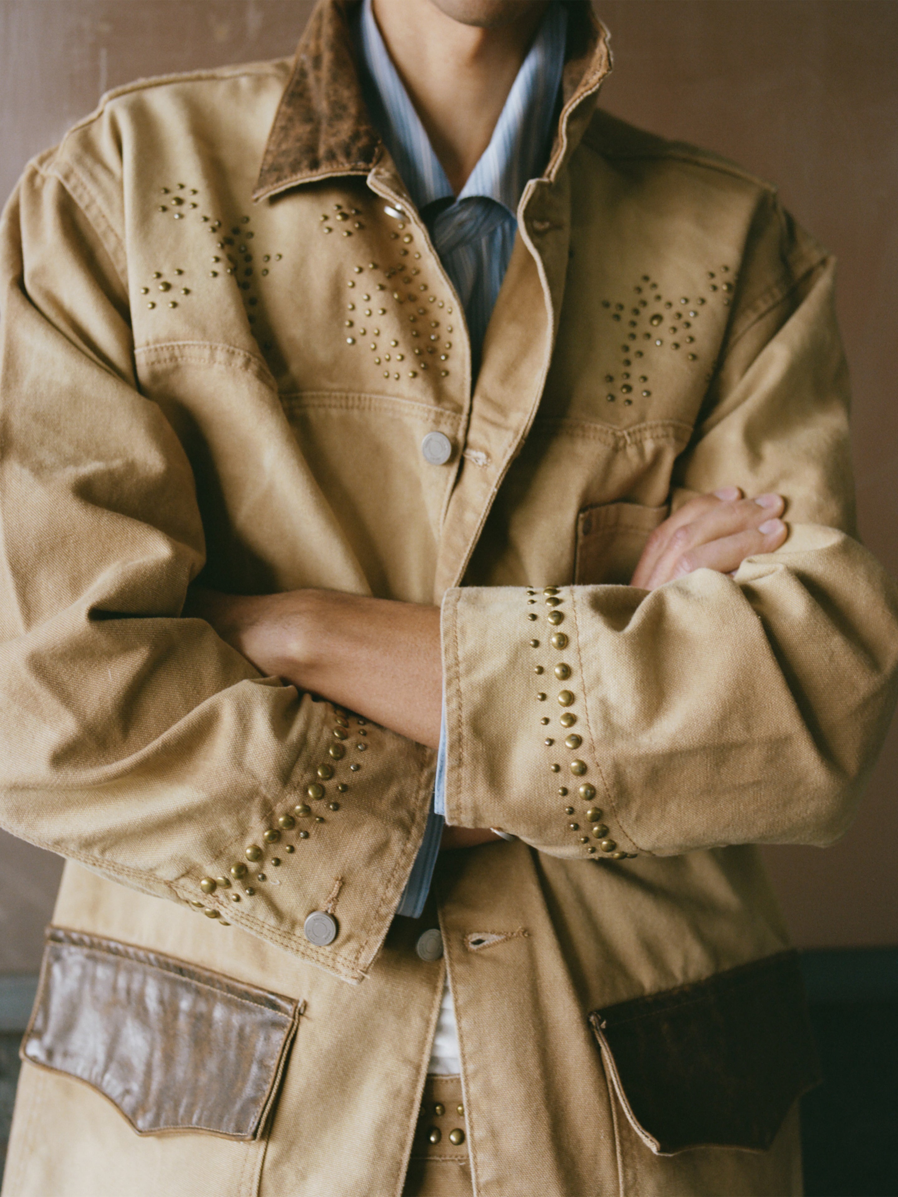 Person wearing the Found Canvas Studded Barn Jacket, featuring metal studs, brown suede accents, and large pockets; arms crossed with face partially out of frame.